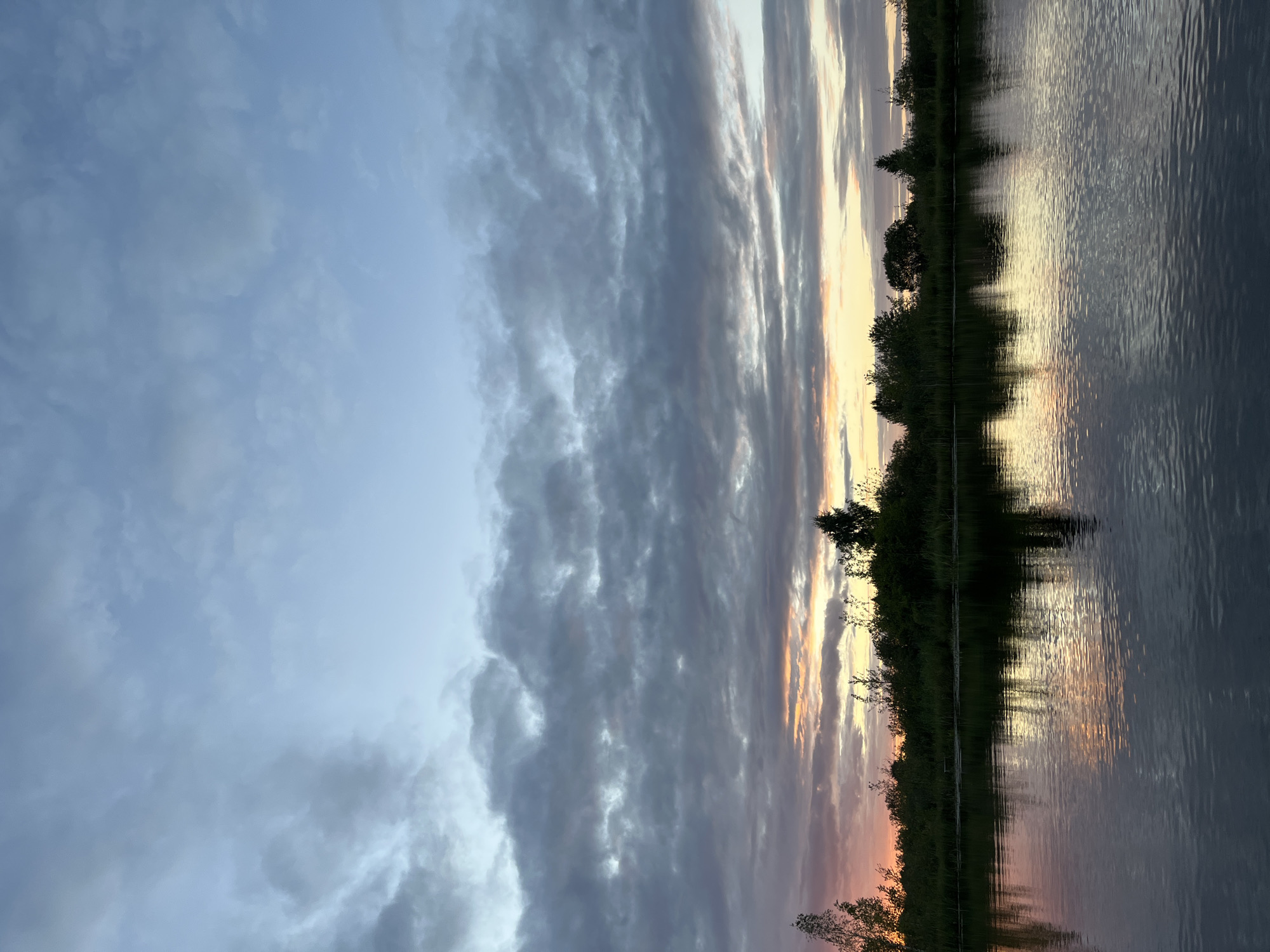 The image depicts a serene landscape with a calm body of water in the foreground, reflecting the sky and the treeline. The sky is filled with a mix of dark and light clouds, with a hint of orange and pink hues suggesting either sunrise or sunset. A dense treeline, composed of various trees and shrubs, forms the horizon. The water appears still, with gentle ripples on its surface. The overall impression is one of tranquility and natural beauty. The specific location is identified as Дунілавіцкі сельскі Савет, Belarus. There are no people or animals visible in the image. The scene is entirely natural and undisturbed.