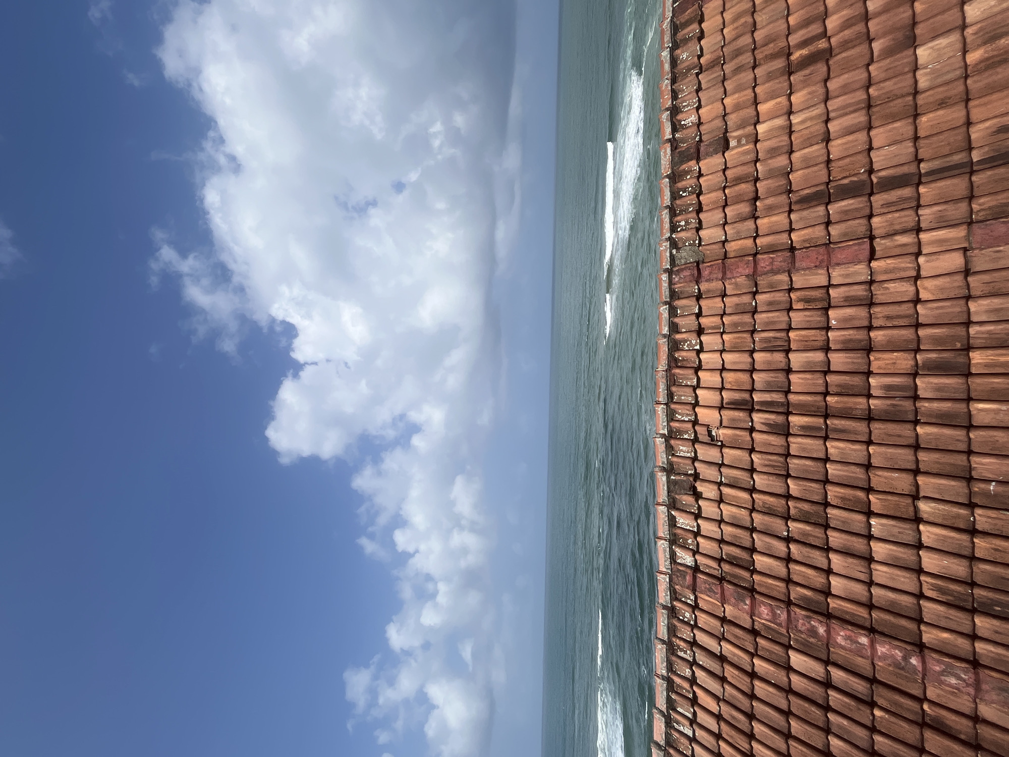 A high-angle shot shows a tiled roof in the foreground, leading to a view of the ocean and sky. The roof is made of weathered, terracotta-colored tiles. The ocean is a deep blue-green, with white waves crashing near the shore. The sky is a vibrant blue, with scattered white clouds. The image appears to be taken from a rooftop in Walgama, Sri Lanka.