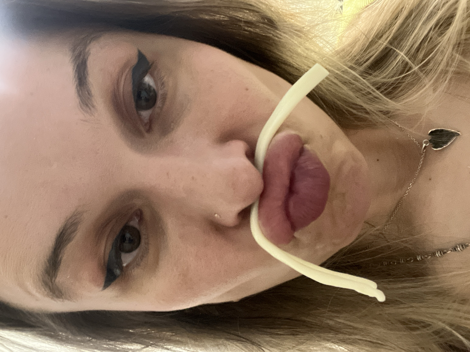 The image is a close-up, vertically oriented photograph of a person's face, with a light-colored, thin object, possibly a piece of pasta or a straw, held between their lips. The person has brown eyes and dark eyebrows, with a prominent winged eyeliner. Their lips are full and painted with a deep pink lipstick. A small nose stud is visible. The person has blonde hair, and a delicate silver necklace with a dark, leaf-shaped pendant rests on their collarbone. The background is blurred, but appears to be indoors with soft lighting. The overall impression is playful or whimsical, suggested by the unusual placement of the object in the mouth. There is no visible text in the image.