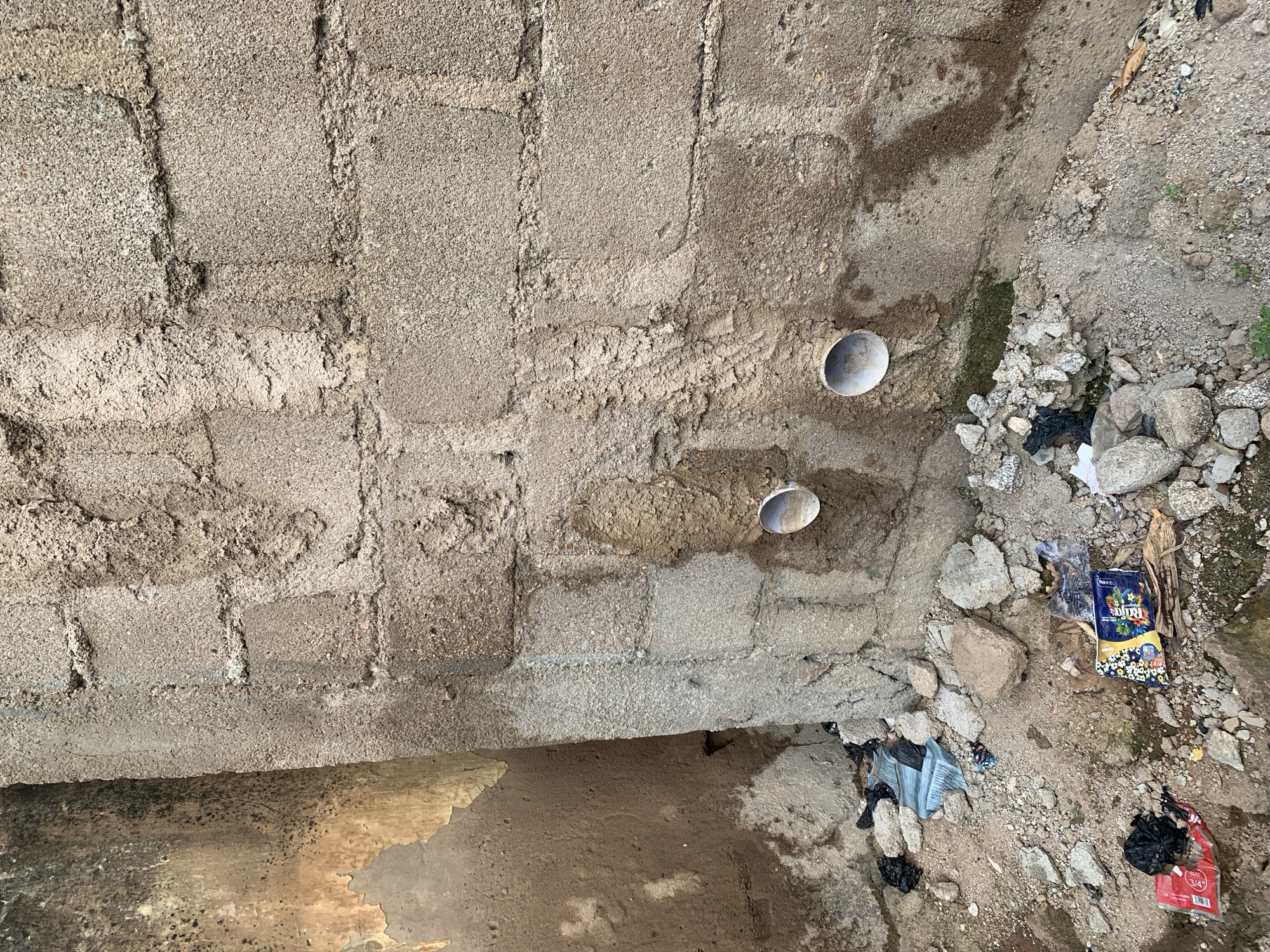 In the city of Unknown, Unknown, a scene of disrepair is captured in this outdoor photograph. The foreground is a rough, uneven terrain composed of dirt, rocks, and scattered debris, including plastic bags, crumpled paper, and what appear to be wrappers. A section of a concrete block wall dominates the mid-ground, showing signs of age and weathering. Two circular pipe openings are visible in the wall, one slightly above and to the left of the other. The wall itself is partially covered in a rough, textured plaster that is stained and discolored in places, suggesting dampness or runoff. To the left of the main wall, another wall is visible, also showing signs of decay with peeling paint and dark discoloration. The overall impression is one of neglect and abandonment.