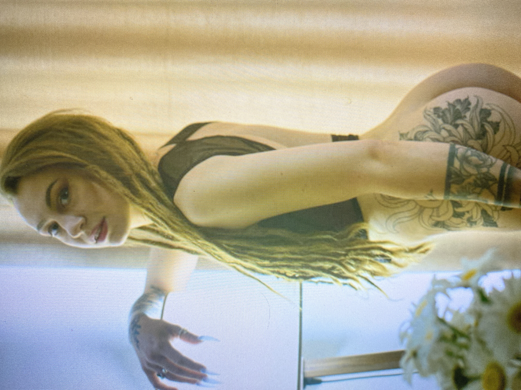 A woman with blonde dreadlocks is posing for a picture. She is wearing a black top and is turned away from the camera, showing her back and buttocks. She has tattoos on her arm and leg. In the foreground, a bouquet of white flowers is visible. The background consists of a light-colored curtain and a window.