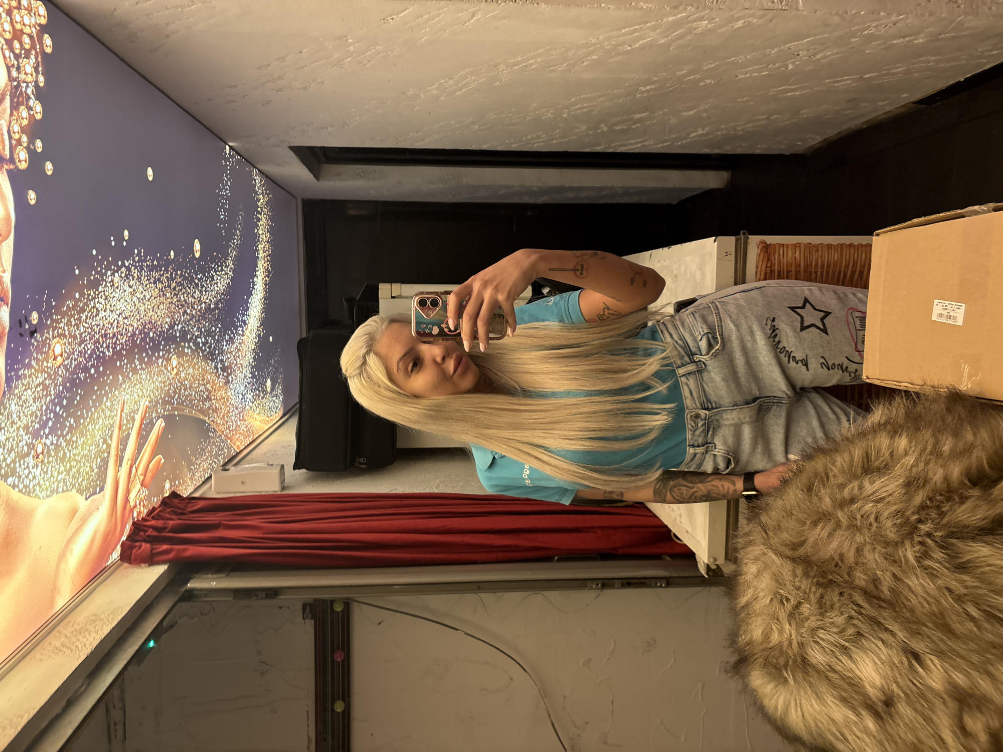 The image, when viewed upright, captures a young woman taking a selfie indoors. She is positioned towards the right side of the frame, holding a smartphone with a decorative case, and looking directly at the camera. She has long, straight blonde hair, wears a light blue short-sleeved t-shirt, and light-wash denim jeans. Tattoos are visible on both her arms, one appearing to depict a chemical structure.
The woman is reflected in a large, vertical display screen or mirror that occupies the left side of the scene. This screen showcases a vibrant digital image: a partial human face (visible from the left, including an eye and nose area) enveloped by a sparkling cascade of golden, jewel-like elements set against a dark background.
The indoor setting appears to be an eclectic space, possibly a commercial area or a room featuring a large digital display. Below the screen, a red, gathered curtain or drapery is visible. In the foreground on the right, there's a large, shaggy brown and tan furry object, resembling a blanket or throw, and behind it, a brown cardboard box with a white, illegible label. The background features a textured light-colored ceiling and a dark horizontal beam. Artificial lighting illuminates the scene.
Visible text includes a partial number "380.3" on her t-shirt sleeve and some illegible script and a black star design on her jeans. The location is Plovdiv, Bulgaria.