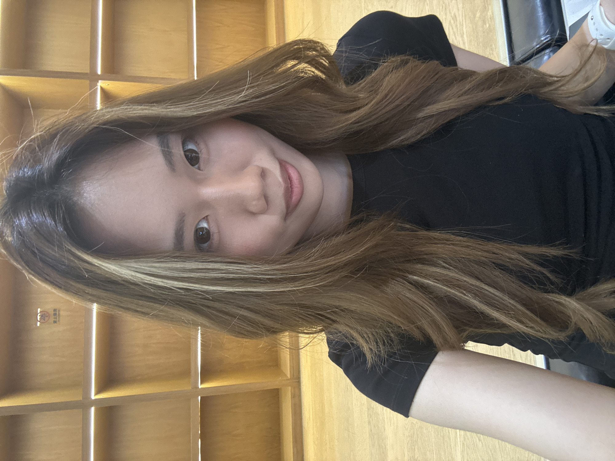 A woman with long, brown hair and a black shirt is taking a selfie indoors. Behind her is a light brown wooden shelving unit. The location is Nan'an District, China, as indicated by a small sign on the shelving unit.