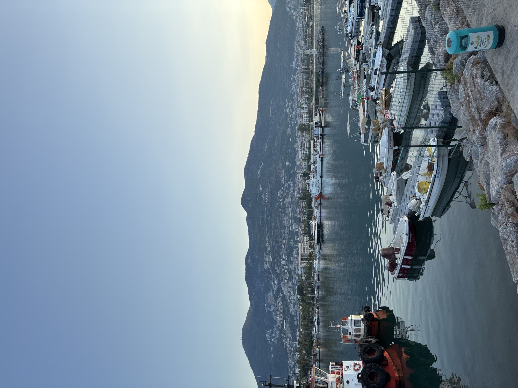 This image captures a serene harbor in Alanya, Turkey, bathed in the soft light of either early morning or late afternoon. In the foreground, several small boats are docked, their reflections shimmering on the calm, deep blue water. One larger tugboat, painted in red and white, is prominently positioned on the left, equipped with large tires for protection. Beyond these boats, a collection of various vessels, including yachts and smaller motorboats, are moored in a marina.
The middle ground reveals the Alanya cityscape, a densely populated area of buildings climbing the hillsides. A prominent mountain range serves as a dramatic backdrop, with its slopes dotted with structures and communications towers. The word "ALANYA" is visible in large letters on the mountainside, serving as a clear identifier of the location. A white lighthouse stands near the harbor's edge, a beacon for the maritime activity.
The sky above is a clear, gradient blue, transitioning to a lighter hue near the horizon where the sun is either rising or setting, casting a subtle golden glow. The environment appears peaceful and orderly, with no visible human activity in this specific snapshot. The overall scene evokes a sense of calm and the picturesque beauty of a coastal city.