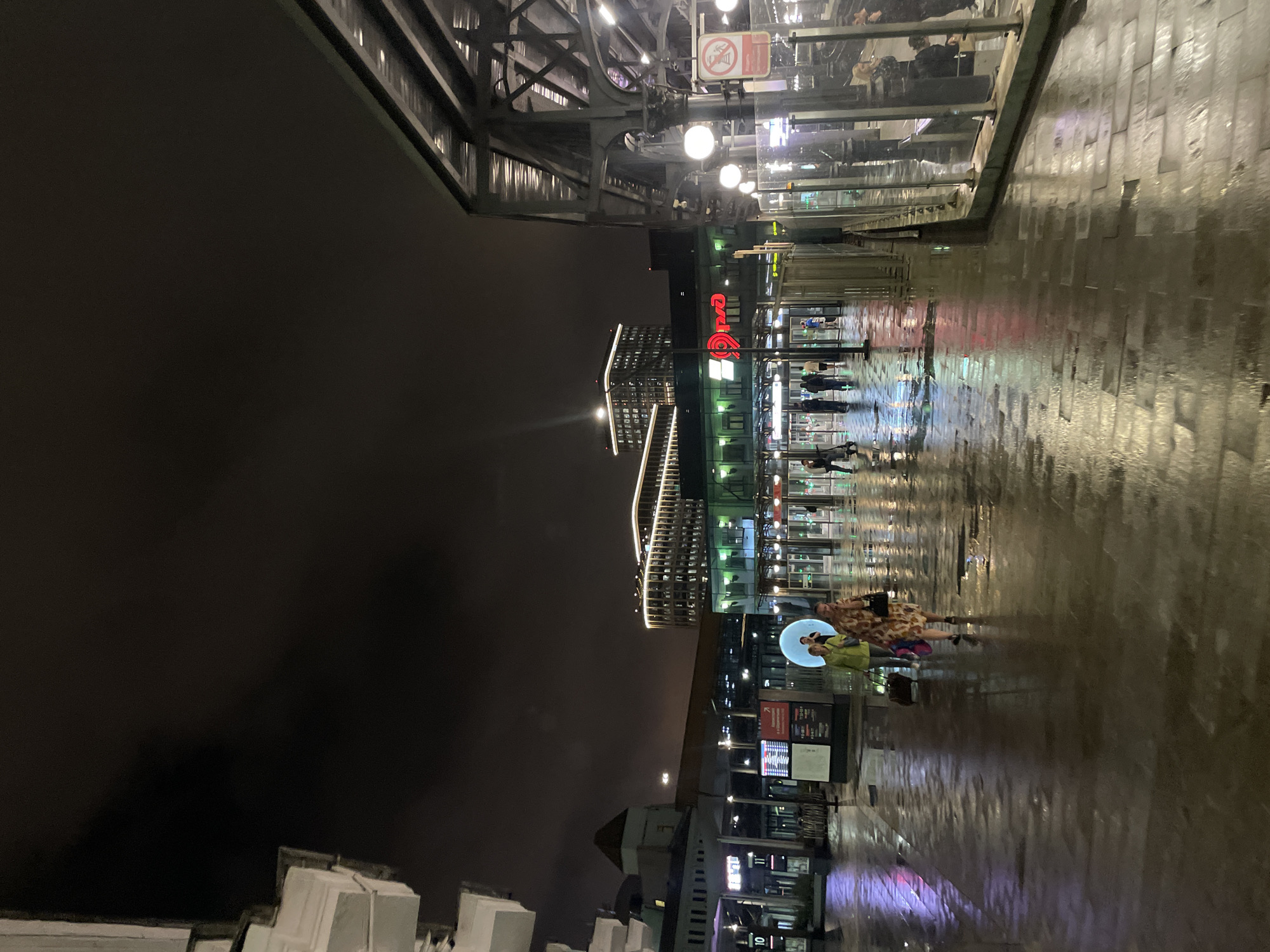 The image captures a night scene outside a train station in Moscow, Russia. The ground is wet and reflective, indicating recent rain, with pavement tiles creating a textured surface. Several people are visible, walking across the wet pavement. In the foreground, two women are walking towards the right, one pulling a suitcase. Further back, other individuals are seen near the entrance of the station.
The station itself is a modern structure, with large glass doors and illuminated signage. The prominent red Cyrillic logo of "РЖД" (Russian Railways) is visible on the building, confirming the location as a railway station. A tall, modern building with many illuminated windows stands in the background, suggesting an urban environment. The sky is dark and overcast. The scene is illuminated by artificial lighting from the station and surrounding buildings, casting reflections on the wet ground. A circular blue light installation is also visible on the pavement, adding a unique visual element to the scene.