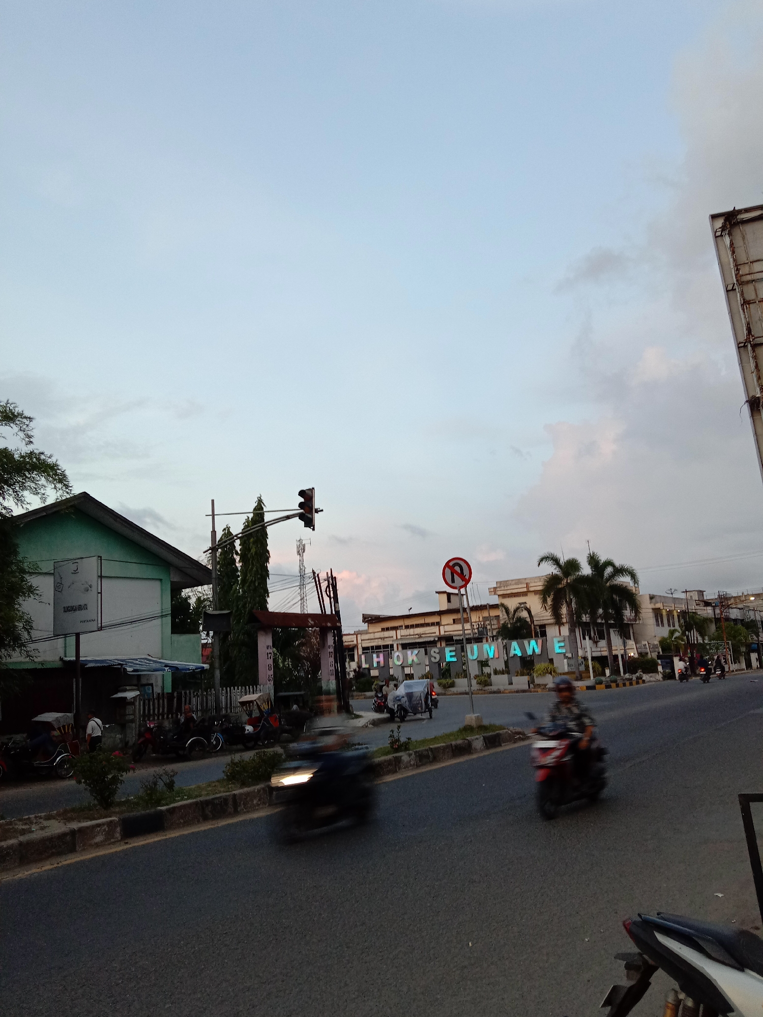 The image depicts a street scene in Lhokseumawe, Indonesia. The street is paved and has a median with some vegetation. There are motorcycles on the road, some in motion, creating blur. On the left side of the street, there is a building with a green facade, and several parked motorcycles or motorbikes near it. A traffic light is visible.
Across the street, there's a building with the illuminated letters "LHOKSEUMAWE" displayed in front of it. A "no U-turn" sign is visible. Palm trees are near this building. The sky is partly cloudy.