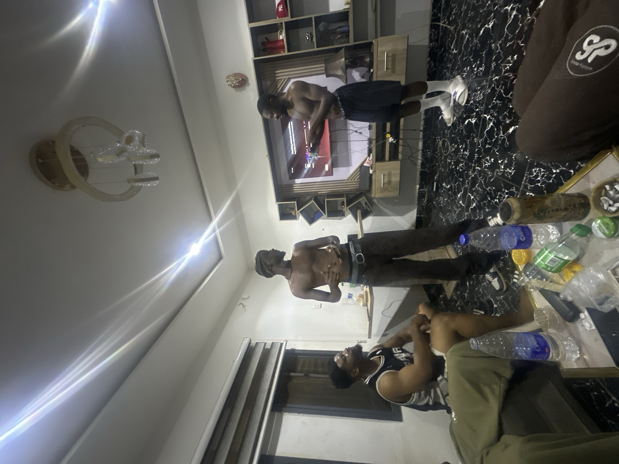 The image captures an indoor scene, likely a living room, where several men are casually gathered. The room features light-colored walls, a dark, reflective floor with a white marble-like pattern, and modern ceiling light fixtures that cast bright reflections.
Three men are distinctly visible. One shirtless man, wearing dark shorts and white sneakers, stands directly in front of a wall-mounted television, appearing focused on its screen. Another shirtless man, wearing dark pants with a belt and a dark head covering, stands further back, looking upwards. A third man is seated or reclining in the foreground, dressed in a dark sleeveless jersey and light-colored pants, also looking upwards. Partially visible in the far right foreground is the arm and leg of a fourth person, wearing brown clothing adorned with a white "SP" logo.
The television is mounted on a wooden entertainment unit that includes shelves and cabinets. The TV screen displays what appears to be a menu or a video game interface with some indistinct text and graphics. On a table in the foreground, several plastic water bottles are scattered, some clearly displaying the brand name "CHAPI". Other items on the table include a brown bottle or can and a small container. The prominent artificial lighting suggests the time of day is evening or night. The context provided indicates the location is Abuja, Nigeria.