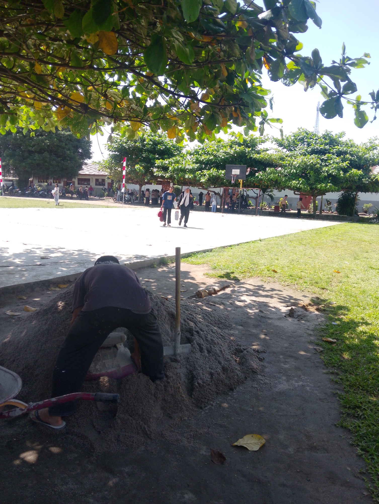 The image depicts an outdoor scene in Serbelawan, Indonesia, on a sunny day. In the foreground, a person wearing a dark purple shirt and dark pants is crouched down, digging into a large pile of sand with a shovel. A cement bag is visible within the pile of sand. To the left of the sand pile, a red-handled shovel and a grey bucket are present. The ground appears to be a mixture of dirt and sand. In the middle ground, a paved area, possibly a basketball court, is visible, with several people walking and standing. Some of these individuals are students, as indicated by their attire. Trees with green and yellowing leaves line the background, with a building and a tall antenna structure visible behind them. The overall environment suggests a school or community area undergoing some form of construction or maintenance.