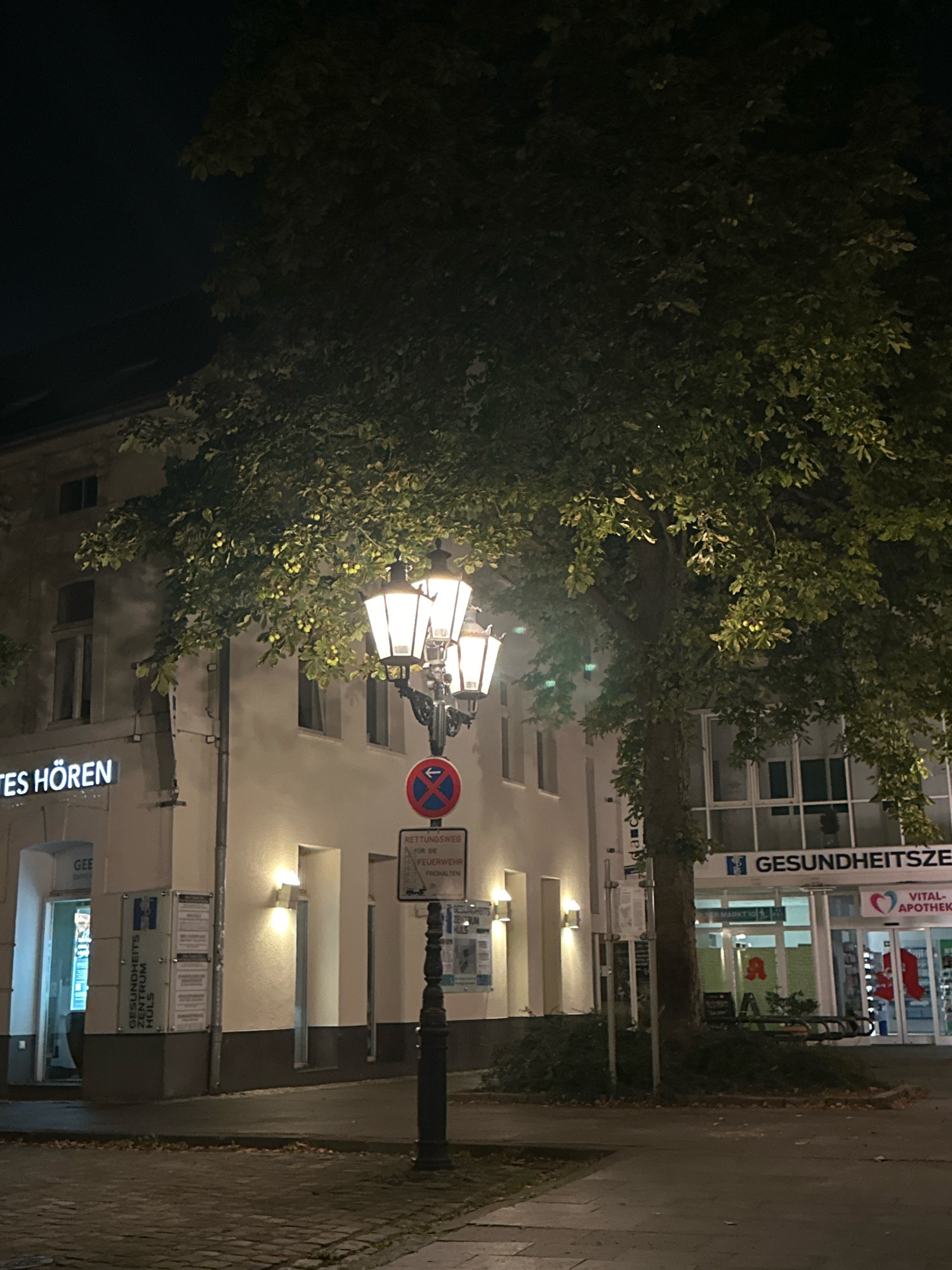 The image depicts a street scene at night in Krefeld, Germany, likely in a commercial or mixed-use area. A large tree with dark green foliage dominates the center and right side of the frame, its branches partially obscuring a building. In the foreground, a black, ornate street lamp with multiple illuminated white lights stands on a paved sidewalk. To the left of the lamp, a sign with red and blue elements indicates a "RETTUNGSWEG" (emergency route) for the fire department.
The building to the left features a lit sign that reads "TES HÖREN". Further to the left, an entrance is visible with a sign that says "GEE" and another with "GESUNDHEITSZENTRUM HULS". The building to the right has large glass windows and displays signs including "GESUNDHEITSZE" (likely part of "GESUNDHEITSZENTRUM") and "VITAL-APOTHEKE". The street and sidewalk are paved with cobbles and asphalt, respectively, and there are some fallen leaves on the ground. The overall lighting is dim, with artificial lights from the buildings and the street lamp creating pools of illumination. No people are clearly visible in the image.
