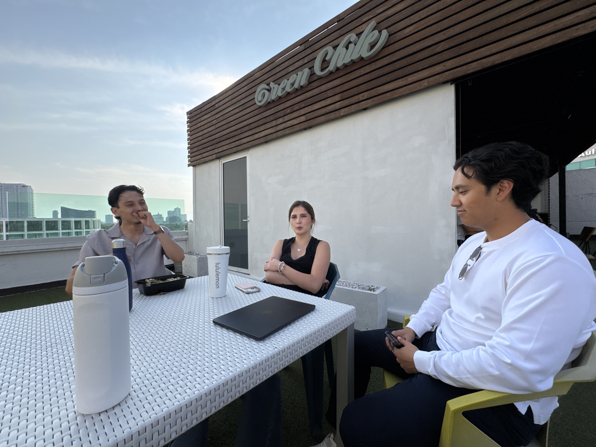 The image shows three individuals—two men and one woman—seated around a white, textured outdoor table, likely on a rooftop patio. The time of day appears to be daytime or late afternoon, indicated by the bright, clear sky with scattered clouds and natural lighting. In the background, a modern cityscape with various buildings is visible.
On the left, a man with dark, curly hair, wearing a light-colored button-up shirt, is smiling and looking towards the left side of the frame. Across the table from him, a woman with long dark hair, dressed in a black top, sits with her arms crossed, looking forward. To the right, another man with dark, curly hair, wearing a white long-sleeved shirt and dark pants, is looking down, holding a smartphone. He has sunglasses hanging from his shirt collar.
Various items are present on the white table: a large white water bottle with a grey lid, a smaller blue bottle, a black take-out container, a white cup with "lululemon" printed on it, a pink smartphone, and a closed black laptop.
Behind the group, a white building wall with horizontal wooden slats above it features a prominent sign in light green script that reads "Green Chile." The setting, with its outdoor seating, distant city views, and restaurant sign, suggests a casual social gathering or meal at an eatery located in Heroica Puebla de Zaragoza, Mexico.