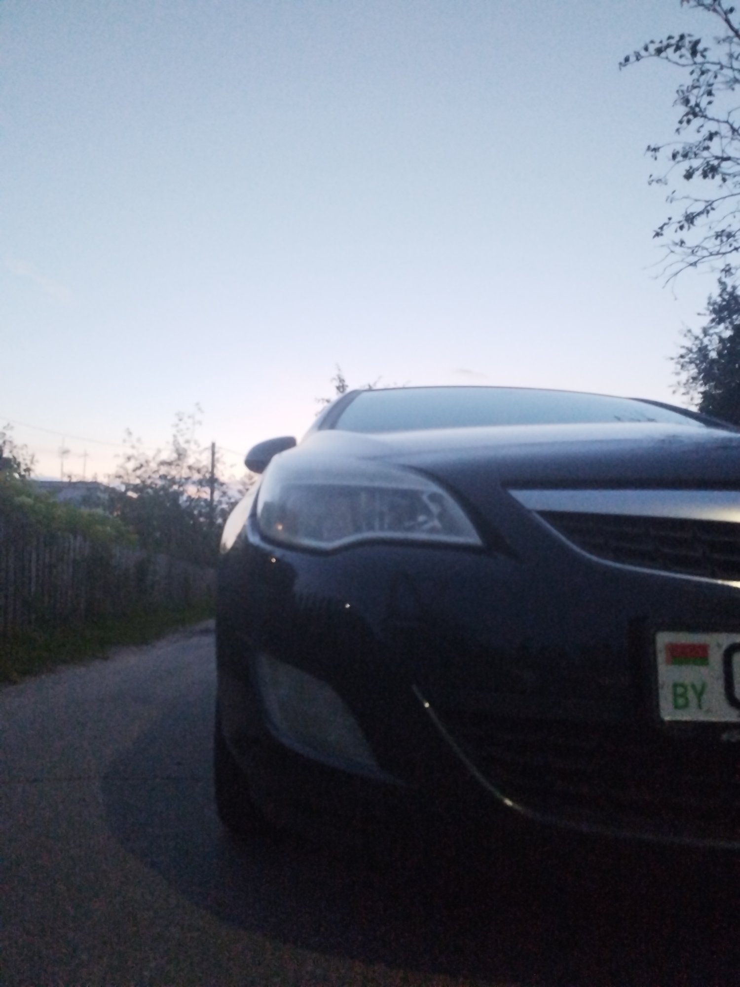 The image, taken at dusk in an outdoor, urban environment, features the front of a dark-colored car. The car is parked on an asphalt road, which is partially visible on the left side of the frame. A wooden fence runs along the left edge of the road, bordering an area with green vegetation. In the background, there are trees and some structures, suggesting a residential or suburban setting. The sky is clear and a light blue, transitioning to a darker hue towards the horizon, indicating the end of daylight. A license plate on the car is partially visible, with the letters "BY" and some other markings. The overall lighting suggests that the photo was taken shortly after sunset.