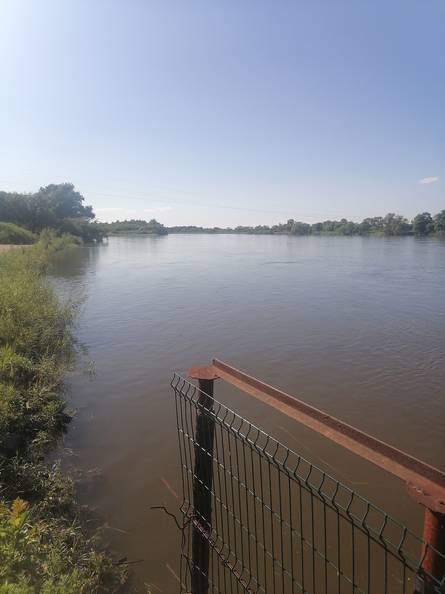 The image depicts a wide, calm river flowing through a rural landscape under a clear, bright blue sky. On the left bank, lush green vegetation and trees are visible, with the bank sloping gently towards the water. The right side of the frame is dominated by the river's surface, which is a murky brown and reflects the sky. In the foreground, a section of a green metal mesh fence with a rusty metal railing runs along the edge of the riverbank, partially obscuring the view of the water. The fence appears to be made of wire mesh panels attached to posts, with a horizontal rusty metal bar on top. The overall impression is one of a serene natural setting, possibly a riverbank or a small dock area. The location is identified as Novopokrovka, Russia. There are no people or animals visible in the image. The scene is static and peaceful.