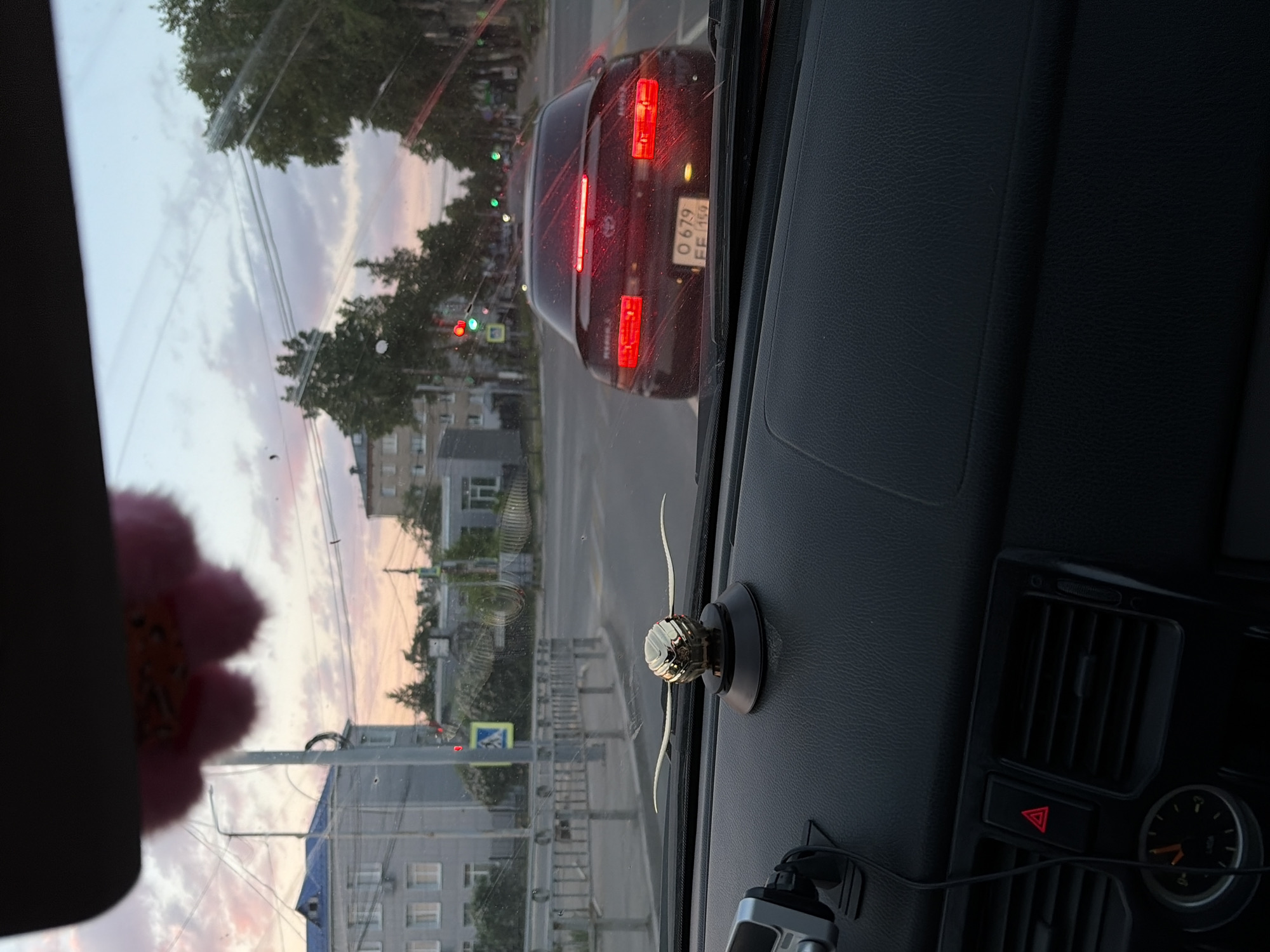 A view from inside a car at a traffic light in Chaikovsky, Russia. Another car with license plate "O 679 EE 159" is stopped ahead. The sky indicates dusk or dawn. A plush object hangs on the left side of the car's windshield, and a decorative item is on the dashboard. The car's hazard lights are activated. Buildings and trees are visible through the windshield.