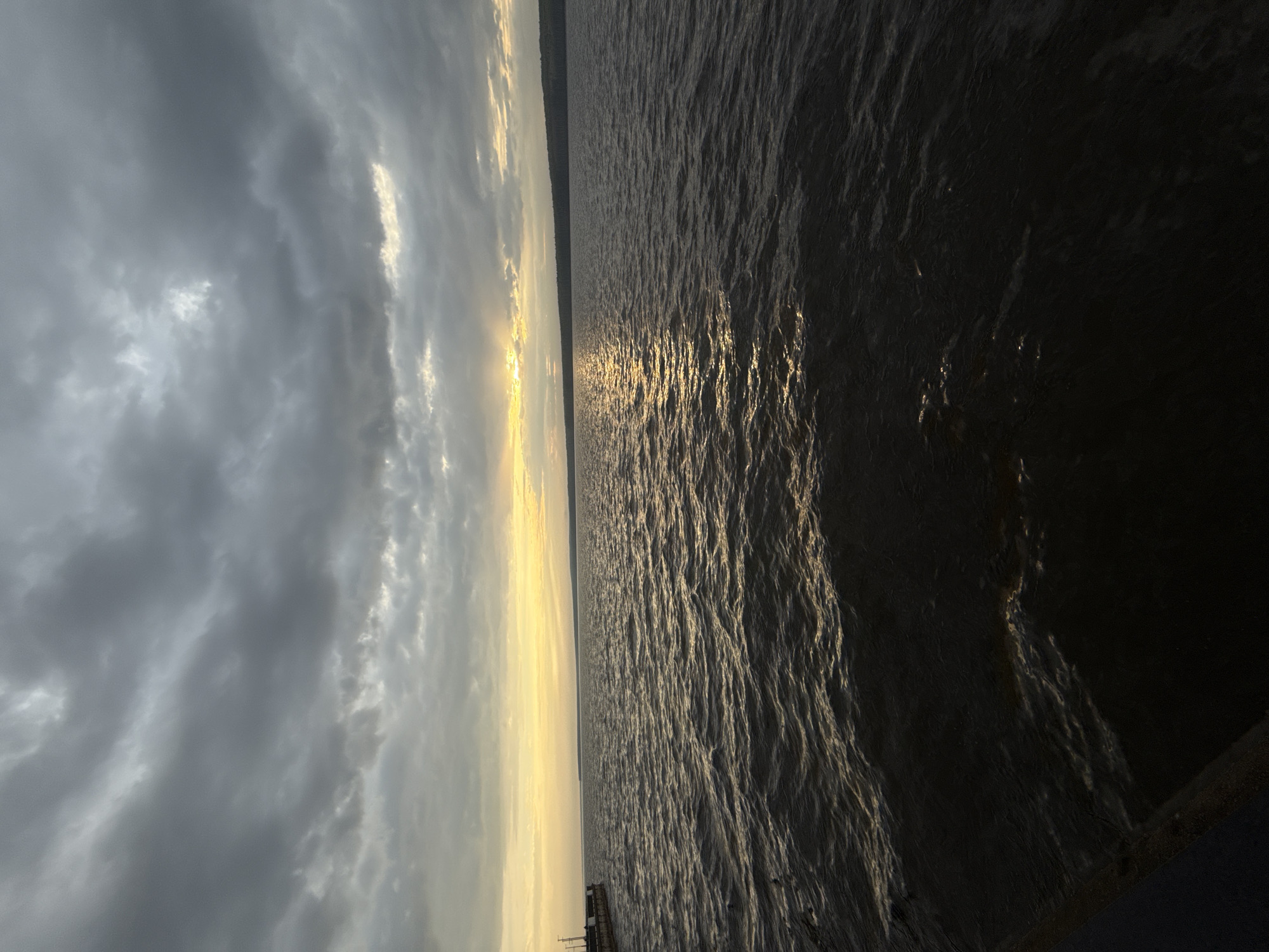 The image shows a body of water, possibly a lake or wide river, under a cloudy sky at sunset or sunrise. The sky is dominated by dark, heavy clouds on the left side of the image, contrasting with a bright, yellow-toned horizon near the center. The sun's rays reflect off the water's surface, creating a shimmering effect. The water itself is dark and textured, with visible ripples and waves. The angle of the photograph is from ground level pointing up towards the sky. The location is likely Chaikovsky, Russia, which is situated on the Kama River, based on the image's characteristics, especially the large body of water. There is a very faint strip of land on the horizon. It is not possible to identify people or actions within the image.