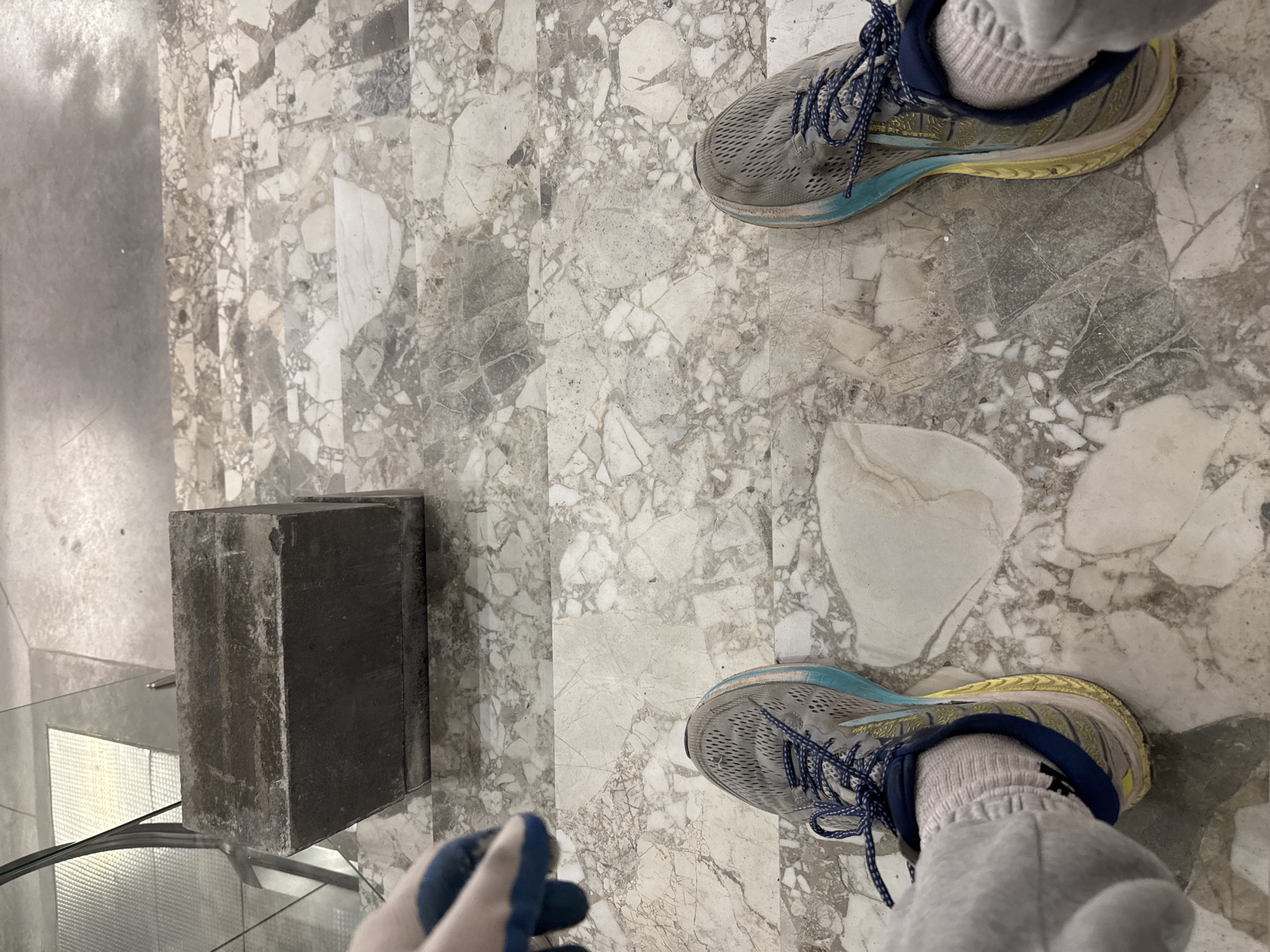 The image provides a first-person, downward view of a person's feet on an indoor floor. The individual is wearing gray sweatpants, white ribbed socks, and a pair of worn grey, blue, and yellow running shoes. Part of the text "PUREFIT SYSTEM" is visible on the side of the right shoe. The person's left hand, adorned with a blue and white work glove, is visible in the lower-left corner.
The floor is made of light-colored, mottled terrazzo or marble tiles, featuring an irregular mosaic pattern of various shades of grey, white, and subtle dark inclusions. These tiles are arranged in distinct rectangular strips that run parallel to each other.
To the left of the feet, a dark, rectangular, block-like object, possibly concrete or heavy stone, sits on the floor. It appears somewhat weathered or dusty. Beneath this block and extending towards the bottom-left edge of the frame is a reflective surface, likely glass or polished metal, which could be part of a display case, a partition, or another architectural element.
The setting is an indoor space, likely a public building or commercial area, characterized by the durable flooring. The lighting is diffused and consistent, suggesting an interior environment. There are no direct visual cues within the image to indicate the provided location context of Kazan, Russia. The scene captures a static moment, focusing on the immediate surroundings of the person's feet, with no explicit activity or emotion discernible.