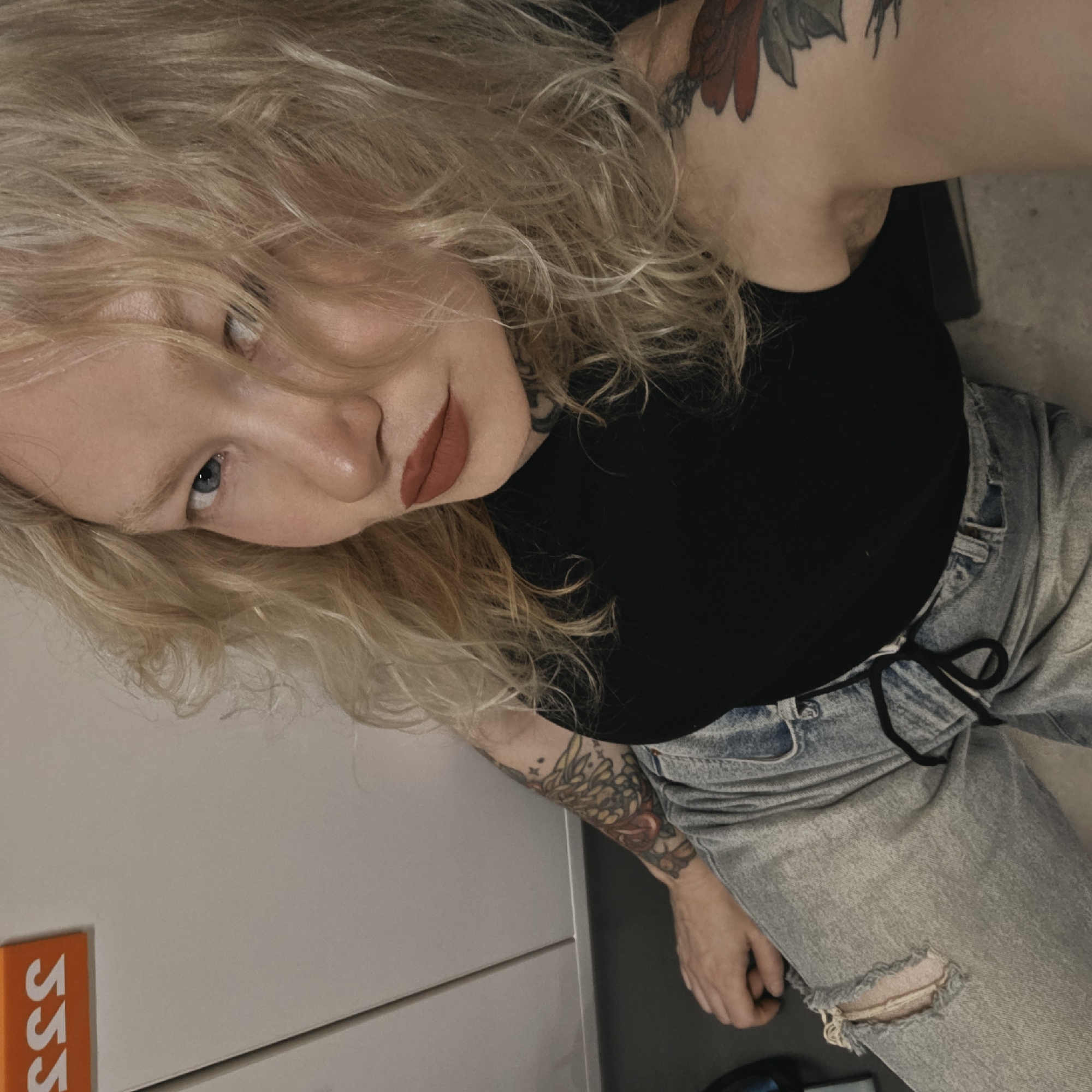 A woman with blonde, wavy hair and light blue eyes is pictured from a slightly upward angle. She is wearing a black top and light blue, ripped jeans with a black drawstring. She has tattoos on her shoulder, neck, and arm. The background appears to be a modern, indoor setting with a white or light gray wall visible to the left. There is an orange sign partially visible on the wall with the letters "SSS." The location in the city of Saint Petersburg, Russia is not discernible from the image.