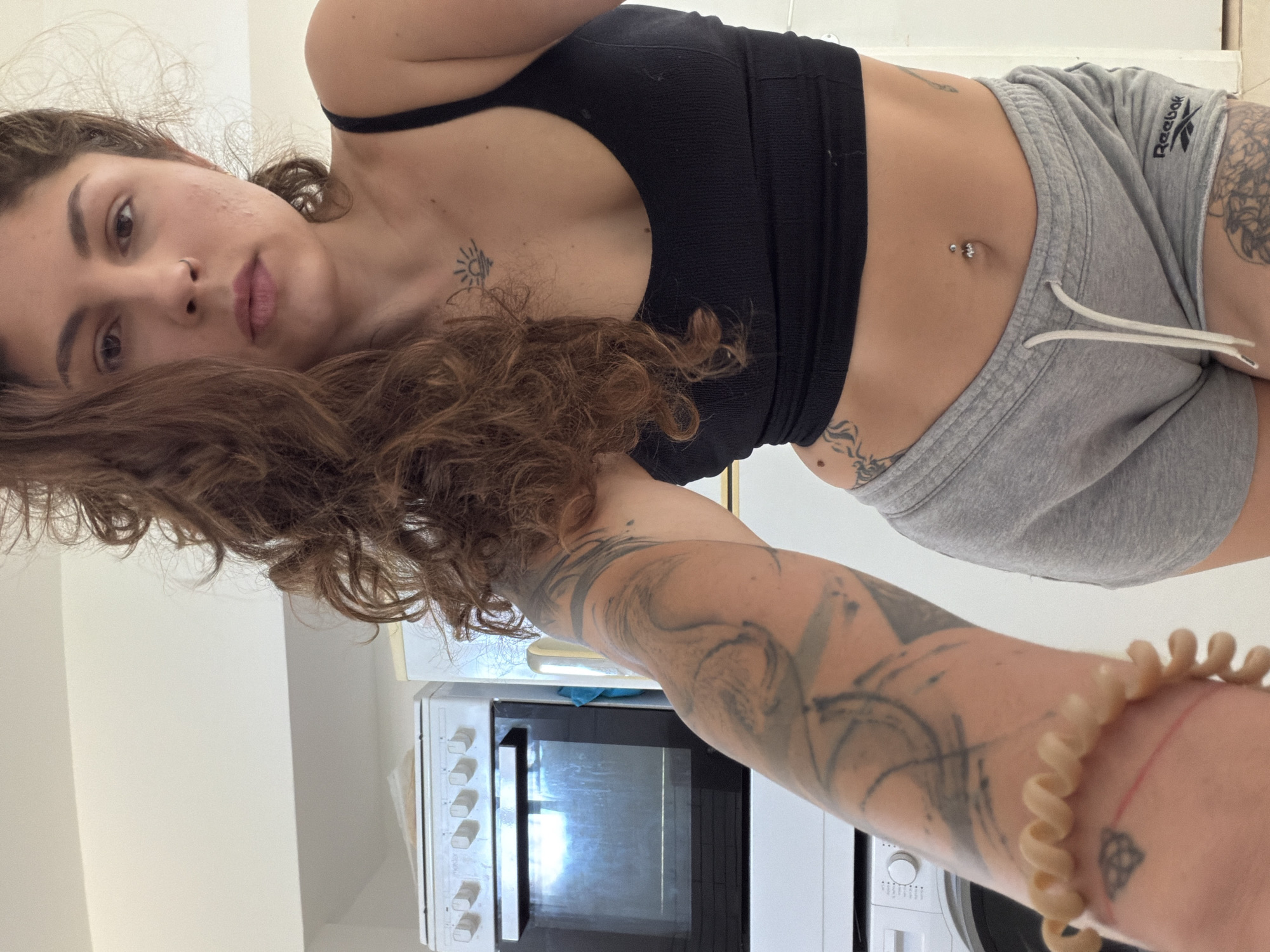 A young woman is captured in a selfie, with the image rotated 90 degrees clockwise, giving the impression she is lying on her side with her head to the left. She has long, wavy brown hair and a neutral expression, looking directly at the camera. Her left cheek shows some visible skin texture. She has a small silver nose stud and a silver belly button piercing adorned with two clear gems.
She is wearing a black ribbed sports bra or crop top and grey Reebok sweat shorts with a white drawstring. Her body features several tattoos: a small stylized sun on her upper chest, extensive abstract or tribal-style designs covering her left arm, and a visible tattoo on her right hip/thigh area. On her right wrist, she wears a light-colored, spiral-shaped hair tie as a bracelet, alongside a thin red string with a small Celtic knot-like symbol.
The background indicates an indoor setting, likely a kitchen or utility room, with a white microwave oven visible on a countertop. The microwave has a dark glass door and several white control knobs. The lighting is bright, suggesting daytime. Her right arm, which appears to be holding the camera, is prominently featured in the foreground, displaying more of her arm tattoos and wrist accessories. The visible text "Reebok" is clearly legible on her shorts. This scene takes place in Kiryat Bialik, Israel.