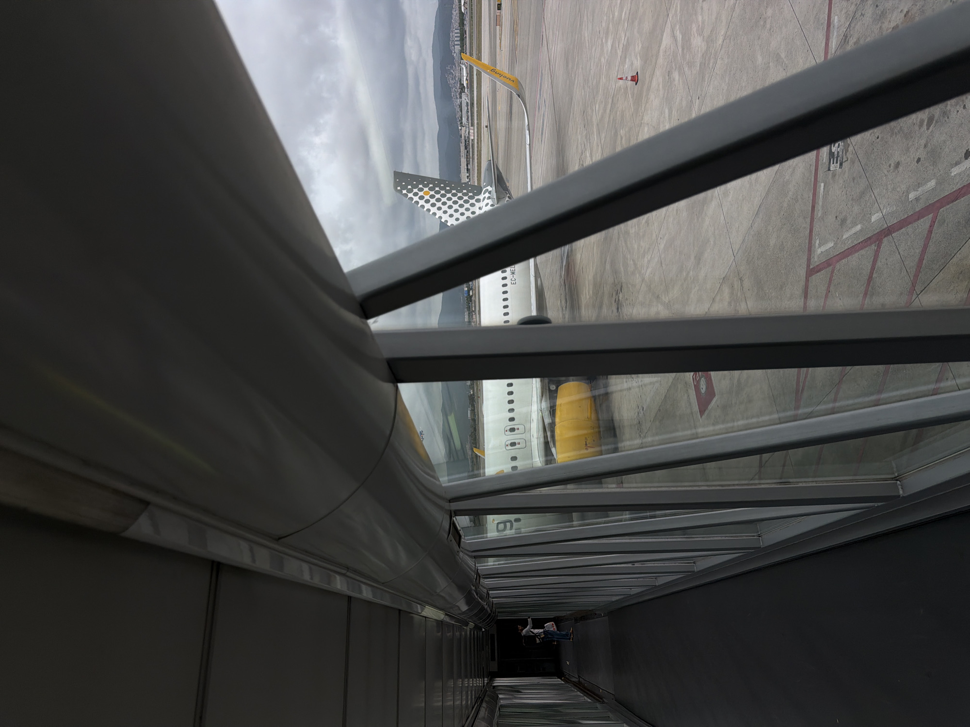 The image, taken from inside a jet bridge, shows a Vueling Airlines airplane parked on the tarmac. The aircraft's tail, with its distinctive polka dot design, and part of its fuselage are visible through the jet bridge windows. The location is likely El Prat de Llobregat, Spain, given that Vueling has a significant presence at Barcelona–El Prat Airport. The tarmac is concrete with painted lines and markings, and an orange cone is visible. A small group of people can be seen walking down the jet bridge toward the plane. The sky is overcast.
