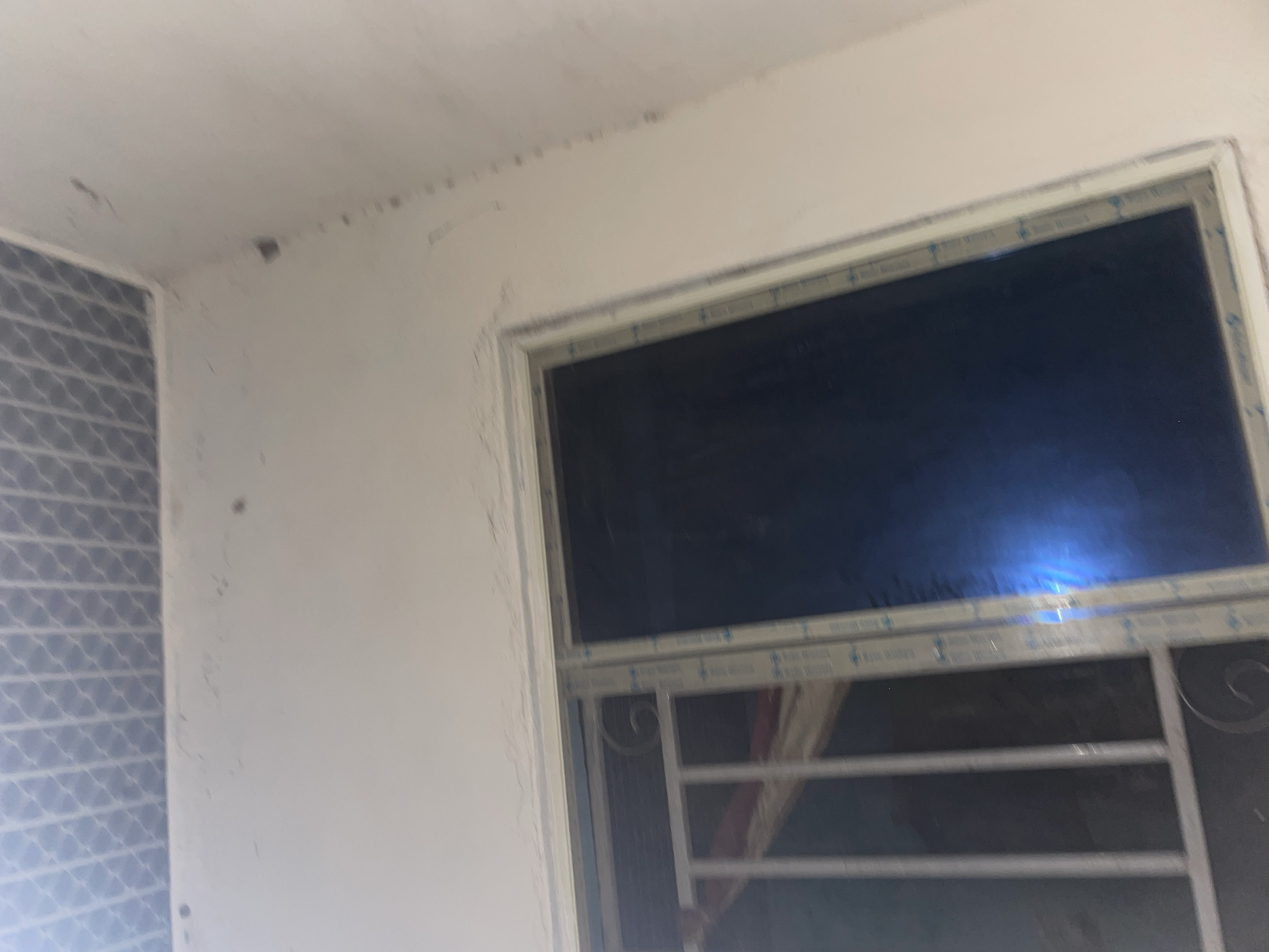 The image shows an interior view of a room in Jalingo, Nigeria, focusing on a window and a section of the ceiling. The ceiling features a patterned white grid, possibly a suspended ceiling or a decorative plaster finish. The walls are painted a light color, showing some signs of wear and tear, including faint marks and what appears to be water damage or discoloration along the top edge near the ceiling.
The window is a sliding type with a white frame. The left side of the window has a white metal grill with decorative scrollwork. The glass pane on the right side is dark and reflective, showing a blueish light source, possibly from an external light or reflection within the room. The window frame itself has some text or branding printed on it in blue.
The overall impression is of a simple, functional interior, likely a residential or commercial space, with some minor maintenance issues visible on the walls. The perspective is angled upwards, suggesting the photo was taken from a low position.