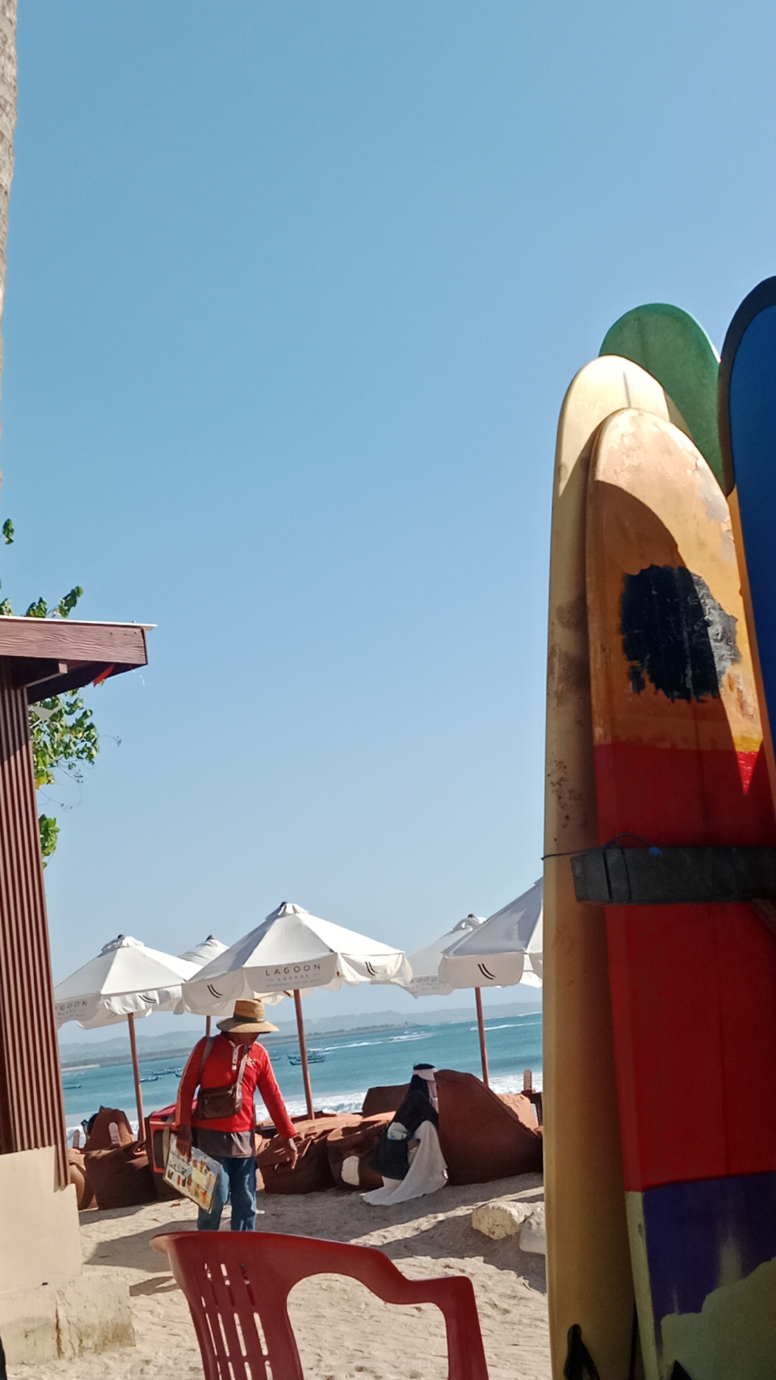 This image captures a sunny day at a beach in Kuta, Indonesia. In the foreground, a red plastic chair is visible on the sand. To the left, a thatched-roof structure with wooden posts provides shade, with some green foliage hanging from it. Further back, several white beach umbrellas with the "Lagoon" logo are set up on the sandy beach. Beyond the umbrellas, the turquoise waters of the ocean are visible, with small boats scattered on the horizon. A person wearing a red shirt, blue jeans, and a straw hat is standing on the beach, holding a magazine. They appear to be a vendor or a beach worker. Next to them, a large beanbag chair is covered with a white cloth, and another person dressed in dark clothing is partially visible sitting or reclining on it. To the right of the frame, several colorful surfboards are leaning against each other, with their fins facing upwards. The sky is clear and blue, indicating fair weather.