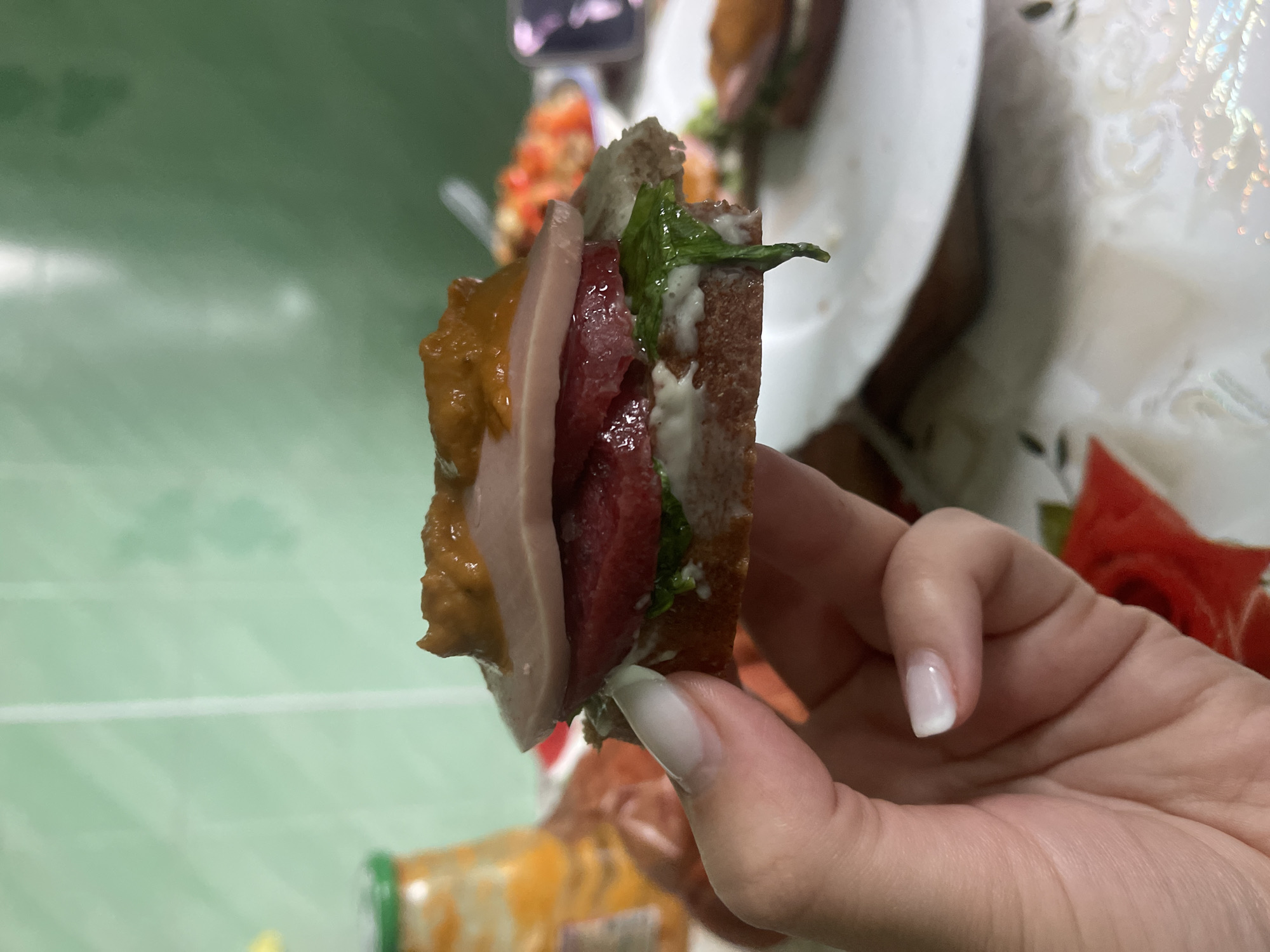A close-up shot shows a hand with well-manicured nails holding a small, layered sandwich. The sandwich consists of a dark bread base, topped with what appears to be white sauce, green leafy vegetables, a slice of red meat, a slice of pale pink meat, and a dollop of a dark, chunky sauce on top. In the blurred background, a glass jar with an orange substance, a plate with food, and a green tiled wall are visible. The image appears to be taken indoors, likely in a kitchen or dining area. The location within the city of Красное, Russia is not discernible from the visual information provided in the image.
