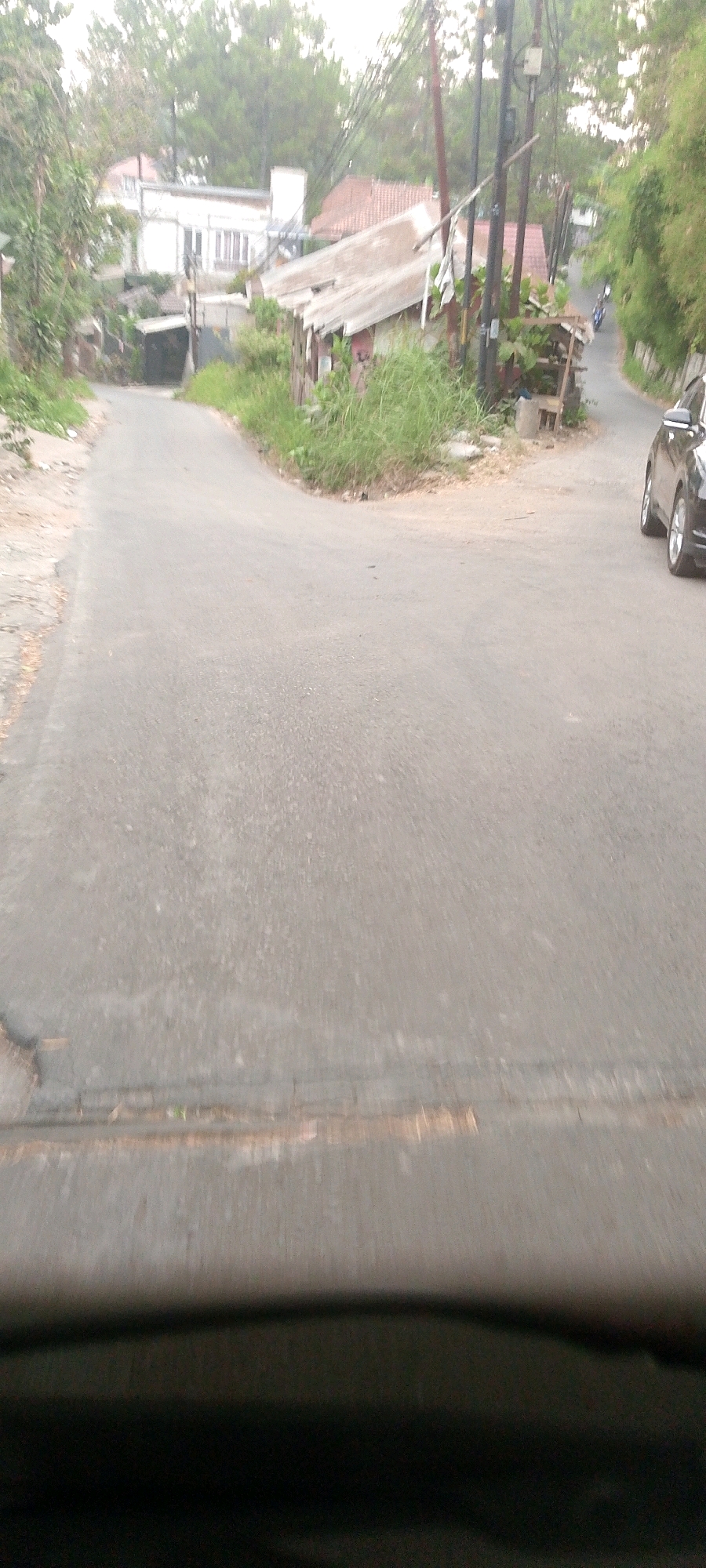 The image depicts a split in a paved road with a slight incline, suggesting an intersection or a fork in the path. On the left, the road continues upwards, bordered by vegetation and a white building in the background. To the right, the road curves and descends, with a dark-colored car visible in the foreground on the right side of the frame. Adjacent to the rightward path, there is a collection of weathered structures, likely makeshift buildings or shacks, with overgrown grass and vegetation surrounding them. Telephone poles with numerous wires are present, indicating utility infrastructure. The overall scene appears to be in a semi-urban or rural environment. The presence of a motorcycle in the distance on the right path suggests it is a public road. The style of the buildings and the vegetation are consistent with some areas in Bandung City, Indonesia, particularly in hilly or less developed regions.