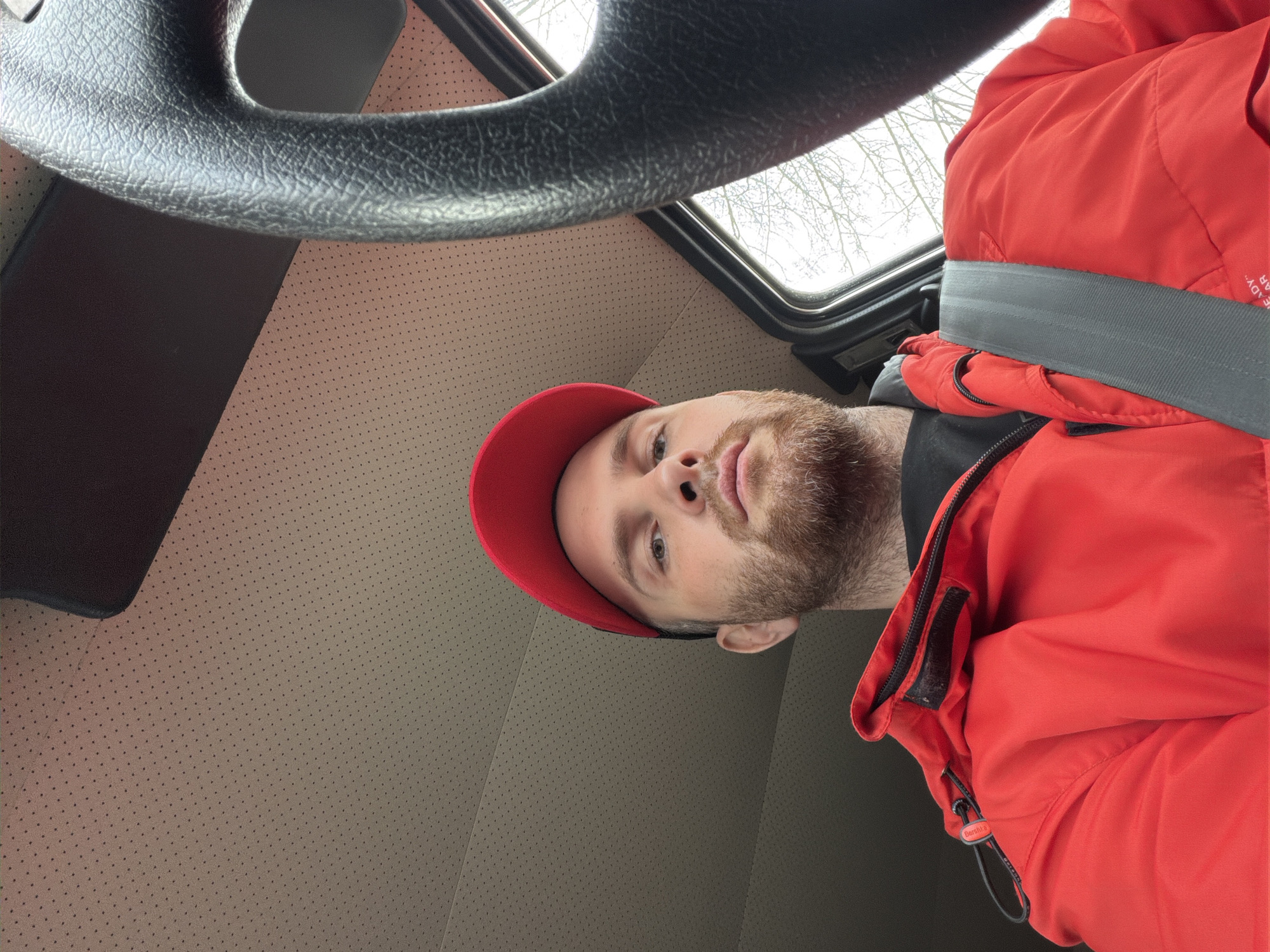 The image is a low-angle selfie taken inside a vehicle, featuring a man wearing a red cap and jacket. The man, positioned towards the right side of the frame, has a beard and is looking directly at the camera with a neutral expression. He is wearing a red baseball cap and a bright red, padded jacket.
The interior of the vehicle is visible, with a dark grey steering wheel occupying the upper left quadrant. The ceiling and upper walls of the car's interior feature a beige headliner with a subtle perforated texture. Through a sunroof or window positioned in the upper center, bare tree branches are visible against a bright, overcast sky, suggesting daytime during winter or early spring.
The man is secured by a grey seatbelt across his chest and shoulder. His red jacket has a black zipper, and a black drawstring with a toggle. On this toggle, the brand name "Berghaus" is clearly visible. A small white tag is also partially visible on the jacket, displaying the word "READY" followed by unreadable text. The setting is a vehicle in Chaikovsky, Russia.