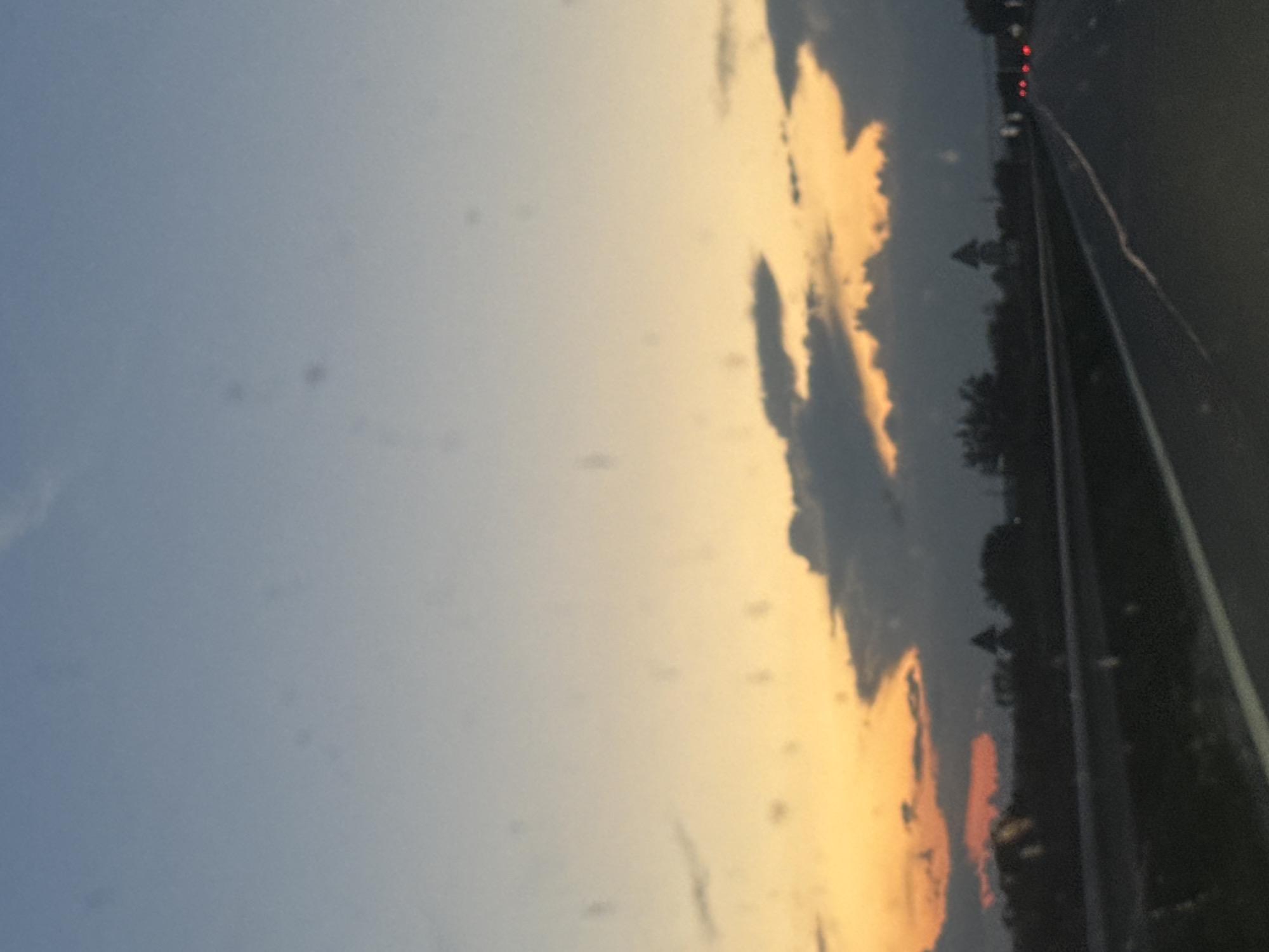 The image captures a twilight scene from a moving vehicle on a highway. The sky displays a gradient of colors, transitioning from a pale blue at the top to warm oranges and yellows near the horizon, indicative of sunset. Dark clouds are scattered across the sky, obscuring some of the lighter hues. Below the sky, a silhouetted landscape is visible, featuring trees and what appear to be structures resembling small towers or steeples, possibly part of the village of Enevo, Bulgaria. The highway itself is in the foreground, with lane markings illuminated by the fading light and distant red taillights of other vehicles suggesting ongoing traffic. The overall impression is one of a journey at dusk, with the natural beauty of the sunset juxtaposed against the man-made infrastructure of the road.