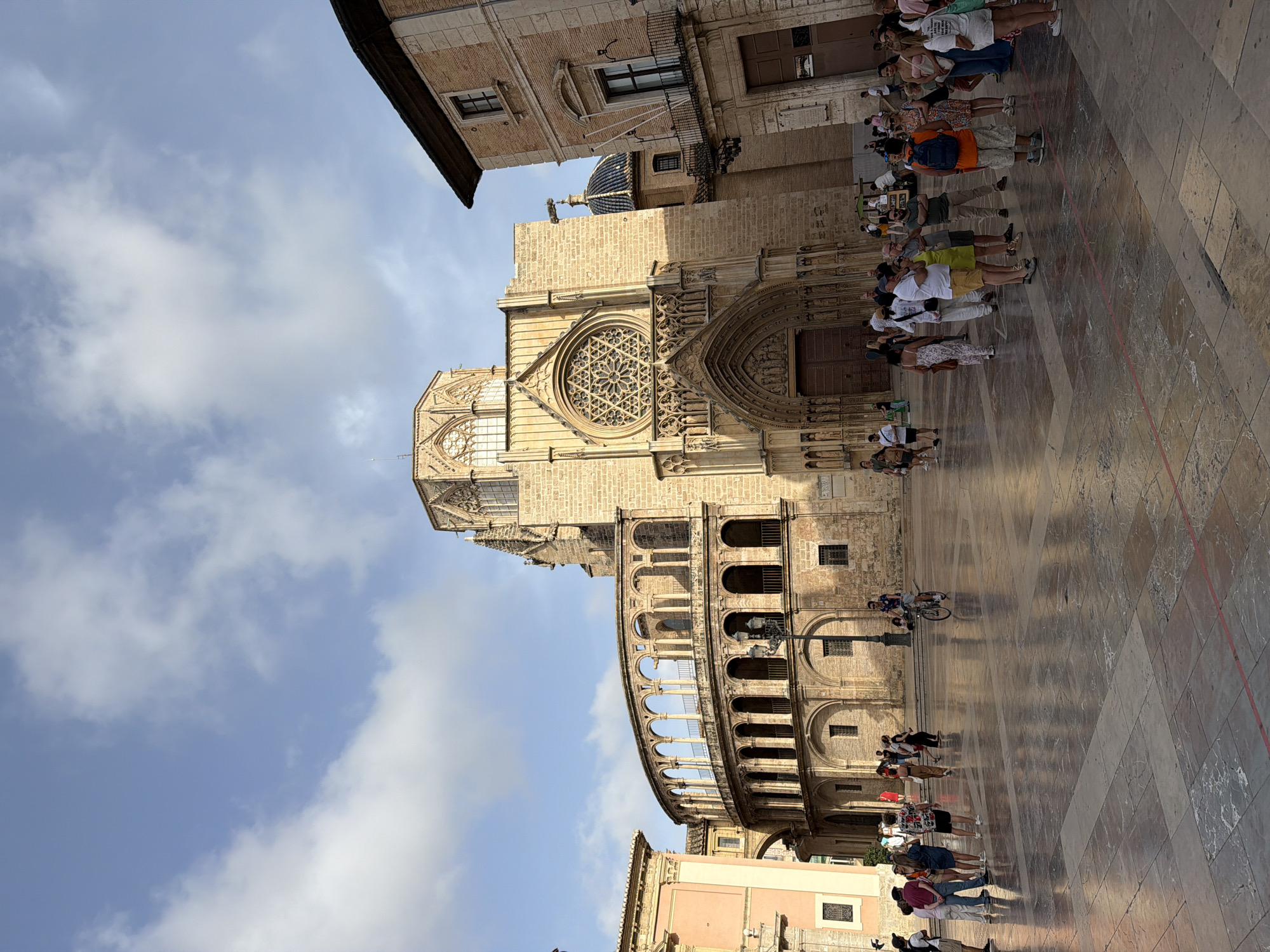 A wide-angle, eye-level shot captures a sun-drenched plaza in front of a grand, historic stone cathedral in an unknown city. The cathedral, with its intricate gothic architecture, features a large rose window and ornate carvings above the main entrance. To the left of the cathedral is a distinctive curved building with multiple arched levels, resembling a cloister or loggia, also constructed of stone. On the right, a brick building with dark shutters and balconies abuts the plaza. The plaza itself is paved with large, polished stone tiles that reflect the sky and surrounding buildings. A crowd of people, appearing to be tourists, are scattered across the plaza, some standing in groups, others walking, and one individual rides a bicycle through the scene. The sky is a clear blue with scattered white clouds. The lighting suggests it is daytime, likely mid-morning or afternoon, with strong sunlight casting shadows.