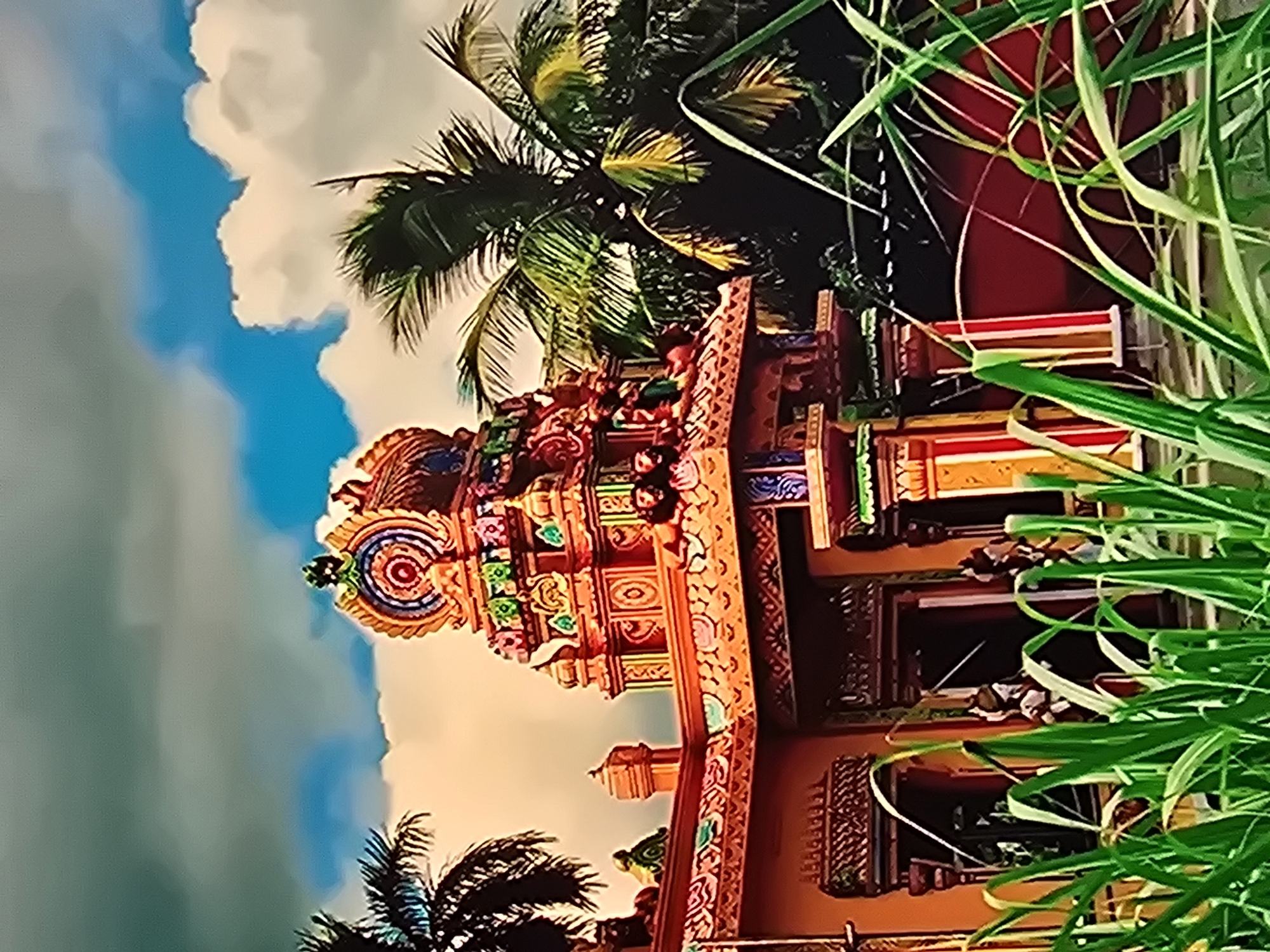 An ornate, brightly colored Hindu temple, likely in Natal, Brazil, is the central subject. The temple features a multi-tiered, intricately decorated tower (Vimana) with statues and carvings. The building's facade is also adorned with vibrant patterns and colors, including red, orange, yellow, and green. Palm trees frame the temple on the left and right, with lush green foliage and tall grass in the foreground partially obscuring the lower portion of the scene. The sky above is a mix of bright blue and dramatic clouds, suggesting a dynamic weather condition. A few figures are visible near the entrance of the temple, though their features are indistinct.