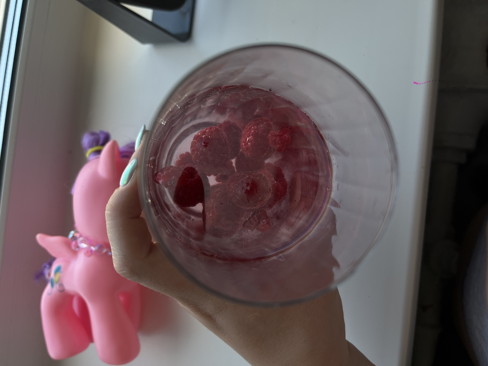 A top-down shot depicts a clear glass filled with raspberries, held in a person's hand. A pink pony toy with a purple mane and tail is visible next to the glass. The background is a white wall with a window frame partially visible in the upper left corner. A dark object is attached to the wall. The lighting is bright and appears to be from natural daylight. There is nothing indicating this is in the city of Izhevsk, Russia.