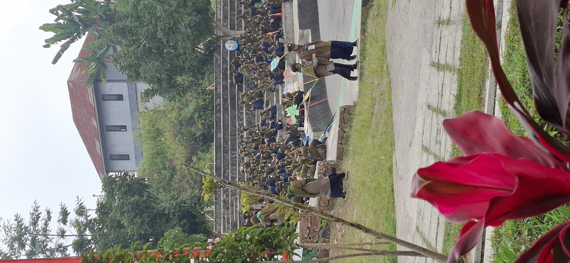 The image captures an outdoor scene, likely in a park or an open-air amphitheater, with tiered seating filled with many people. The majority of the individuals are wearing uniforms, predominantly dark green tops with navy blue bottoms and headscarves, suggesting a group activity or event, possibly related to scouting or a school function. In the foreground, two individuals are walking towards the right, also in similar uniforms. The background features a modern building with a red-tiled roof and grey walls, surrounded by lush green trees and foliage. The overall atmosphere appears to be one of gathering and shared activity. The location is identified as Karang, Indonesia.