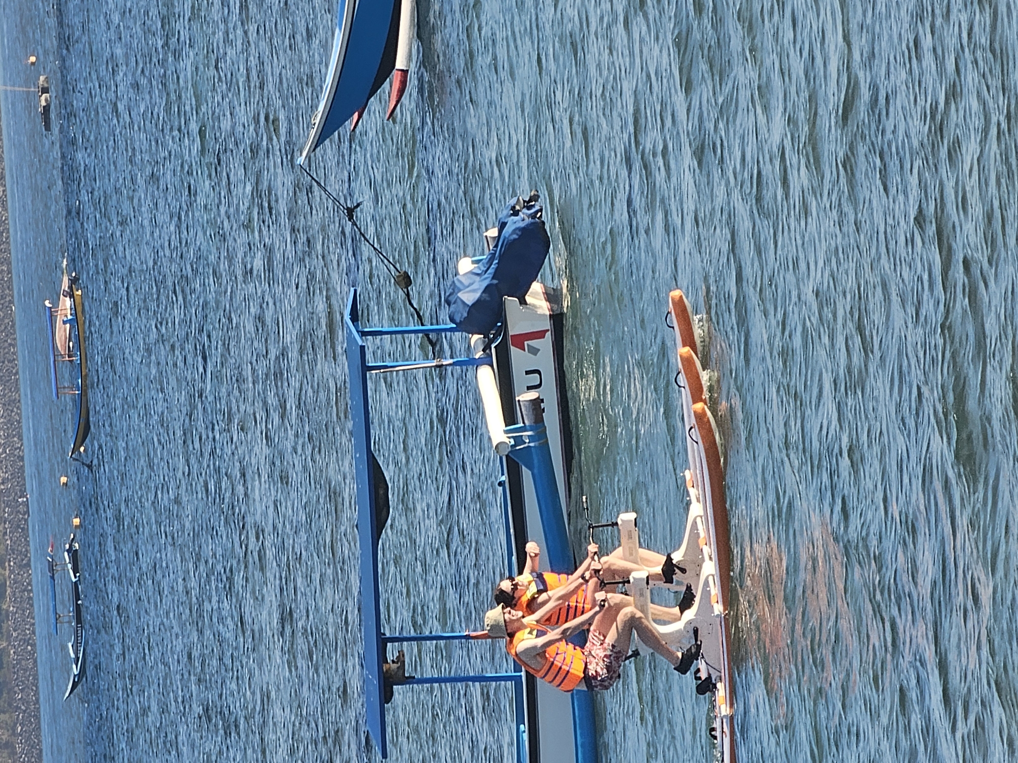 Two individuals, a male and a female, are engaged in riding a water bike on a body of water. The male, on the left, is wearing a light-colored bucket hat, sunglasses, and an orange life vest over patterned shorts. The female, to his right, is wearing sunglasses and an orange life vest. Both are seated and pedaling the water bike, which is white with orange pontoons. In the background, a blue and white boat with a canopy is visible, and further in the distance, several other boats are scattered across the water. The environment appears to be a sunny day with calm water, typical of a coastal area. This scene likely takes place in Denpasar, Indonesia, given the presence of traditional boats and the coastal environment.