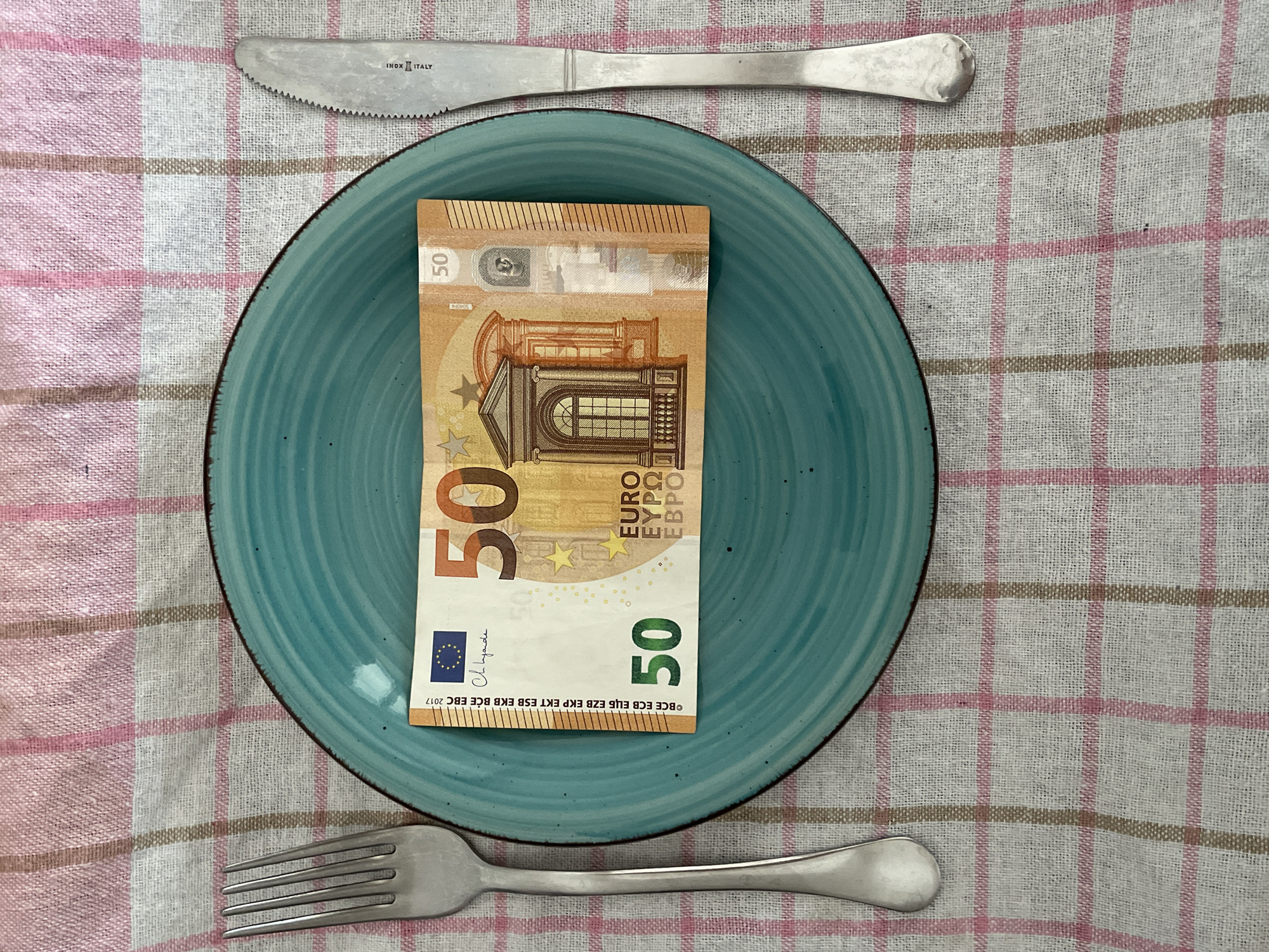 This image captures a table setting from an overhead perspective, situated in Budva, Montenegro. The scene is set on a tablecloth with a prominent white, pink, and light brown checkered pattern. A round, teal-colored ceramic plate, featuring a dark rim and subtle concentric grooves, is centrally placed. To the left of the plate lies a silver-colored metal fork. To the right, a silver-colored metal knife with a serrated blade, bearing the inscription 'INOX ITALY', is positioned. A single 50 Euro banknote, from the 2017 Europa series, is laid horizontally across the center of the plate. No individuals, actions, or interactions are depicted beyond the static arrangement of these items.