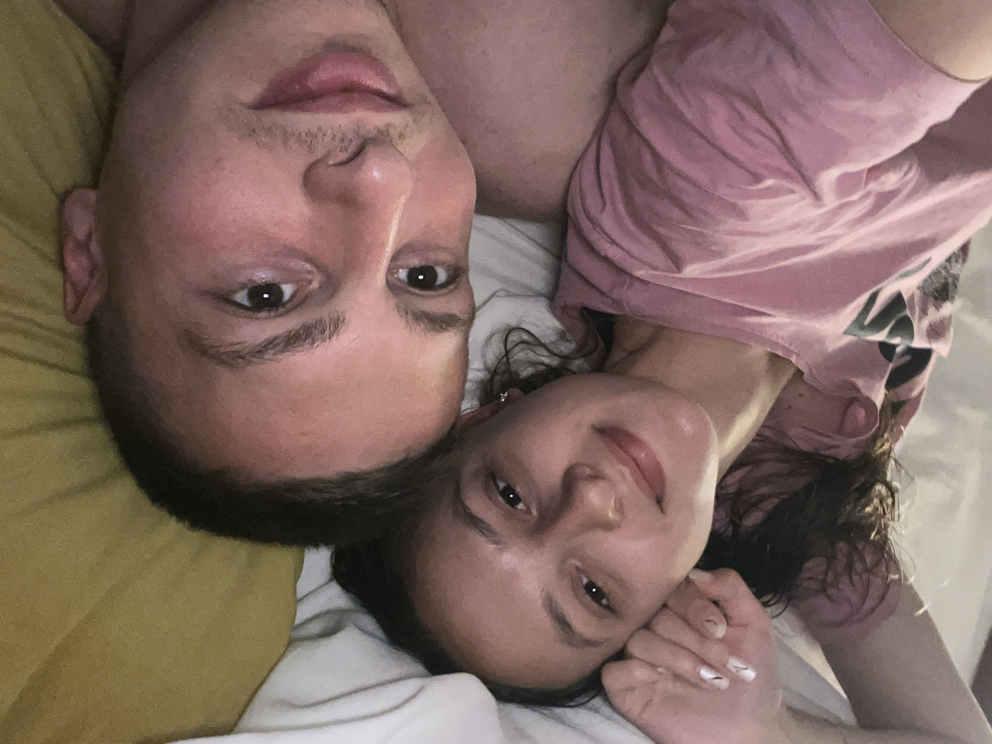 A selfie of two people lying in bed. The man is at the top of the frame, with short hair, and fair skin. The woman is positioned at the bottom, with dark curly hair and fair skin. Both are looking into the camera. The man is shirtless, while the woman is wearing a pink t-shirt. They are lying on white sheets with a yellow pillow visible behind the man's head. The location is Vitry-sur-Seine, France, which is not discernible from the image.