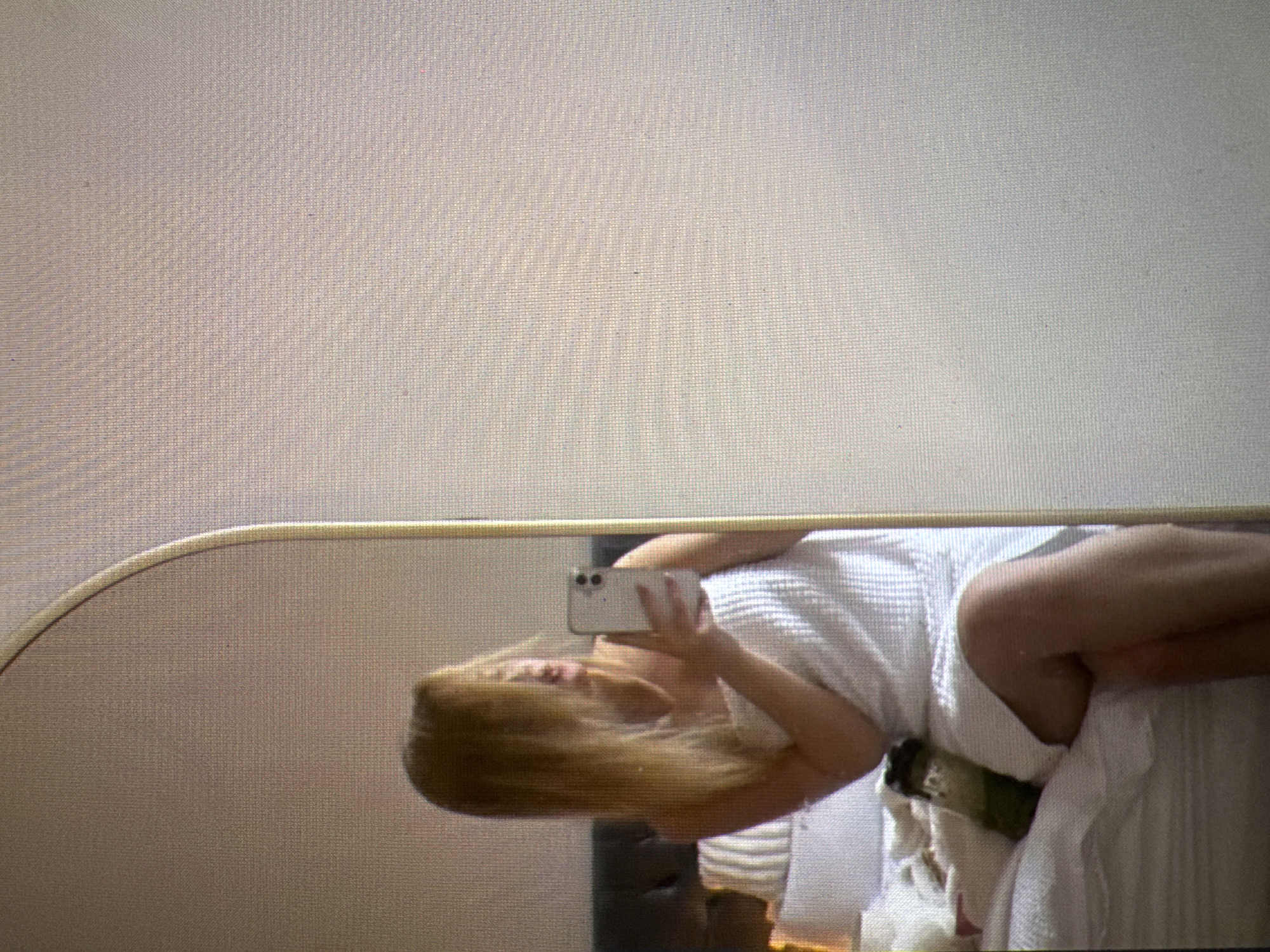 In a bedroom in Castelo Branco, Portugal, a person is captured in a mirror selfie. They are seated on a bed, wrapped in a white towel, and holding a smartphone. The room is lightly furnished, with pillows visible on the bed and a dark headboard. A water bottle with a green liquid is also on the bed. The lighting is soft, and the overall scene suggests a casual, private moment.