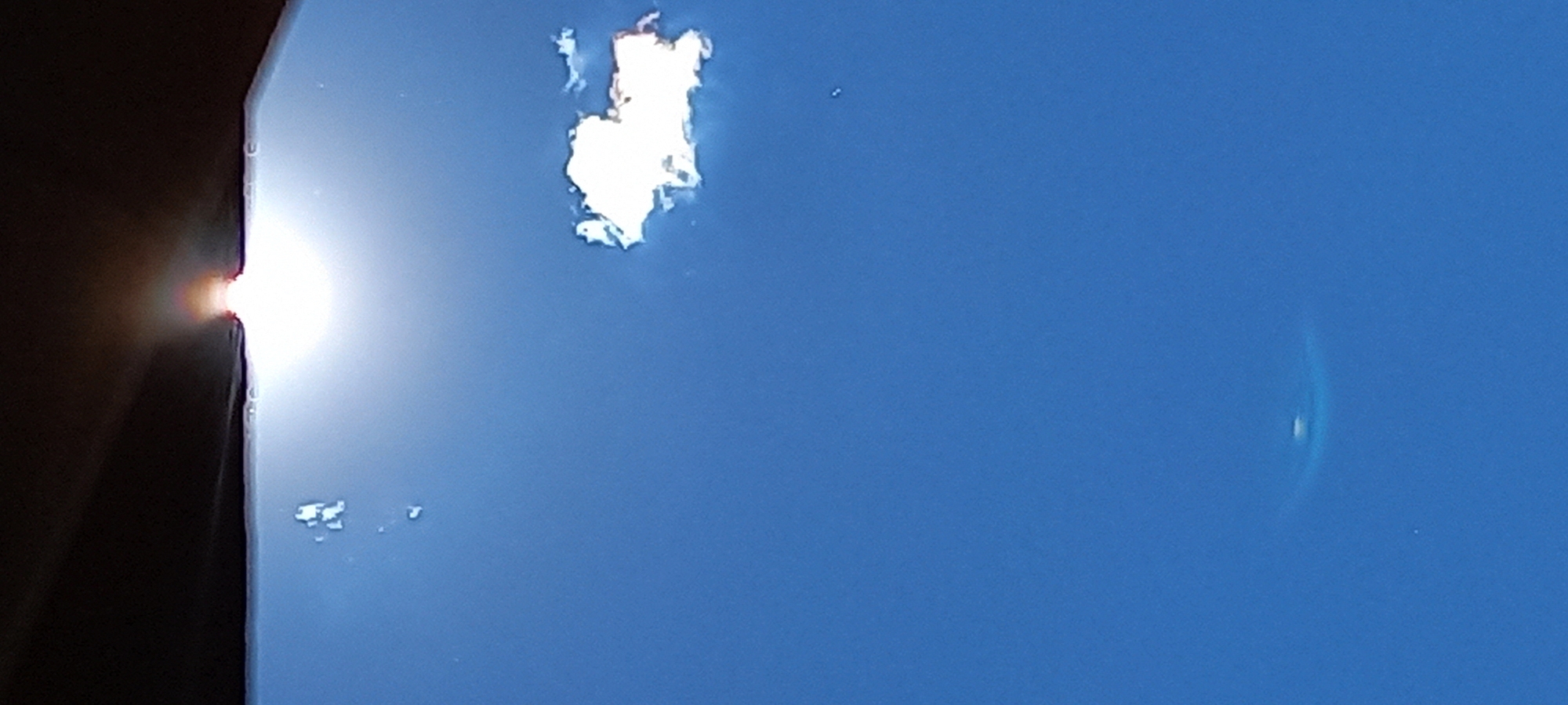 The image shows a section of clear blue sky. In the upper center, there is a bright, irregular patch of light, possibly a reflection or glare. To the left of the frame, there is a dark, out-of-focus object, possibly a building edge, with a bright light source partially obscured by the edge. In the lower-right, a faint circular or oval distortion appears. There is no visible evidence of the location being Kryvyi Rih, Ukraine, nor the presence of people or interactions.