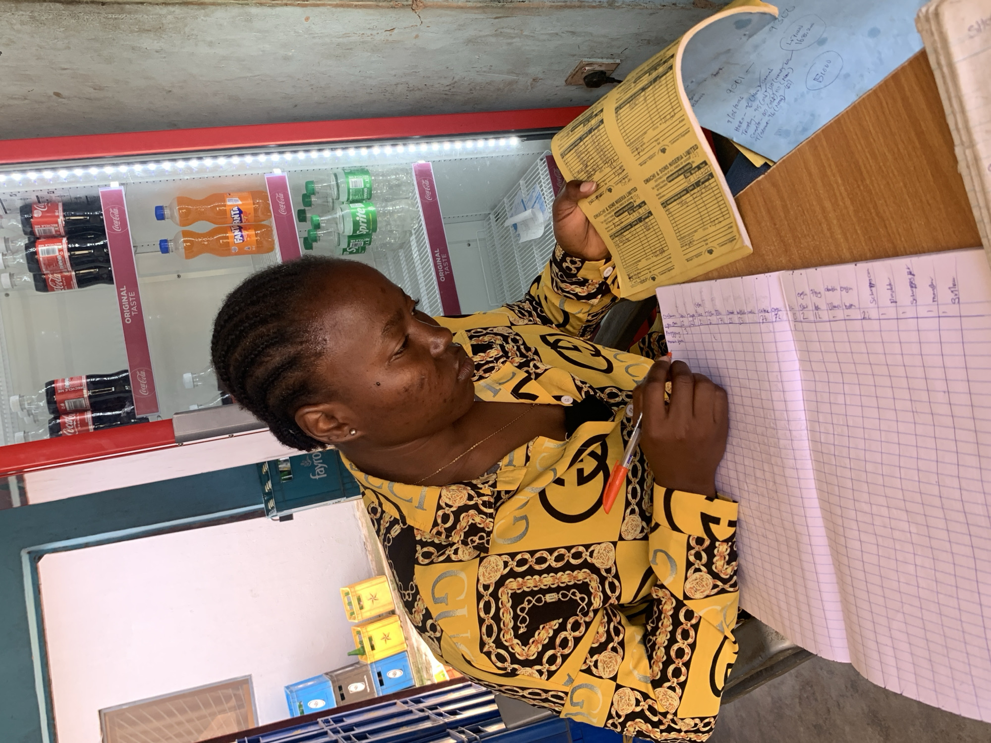 In a retail establishment in Jos, Nigeria, a Black woman is seated at a wooden desk, diligently writing in a ruled notebook. She is wearing a yellow long-sleeved shirt with a black and gold chain print. Her hair is styled in braids. In front of her, on the desk, are the open notebook, a yellow ledger with printed columns, and a light blue paper with handwritten notes. To her right, an illuminated red beverage cooler is stocked with bottled drinks, including Coca-Cola, Fanta, and Sprite. The background shows shelves with various crates of beverages, and a wall with a power outlet. The woman's focus is on her writing, with her right hand holding a pen.