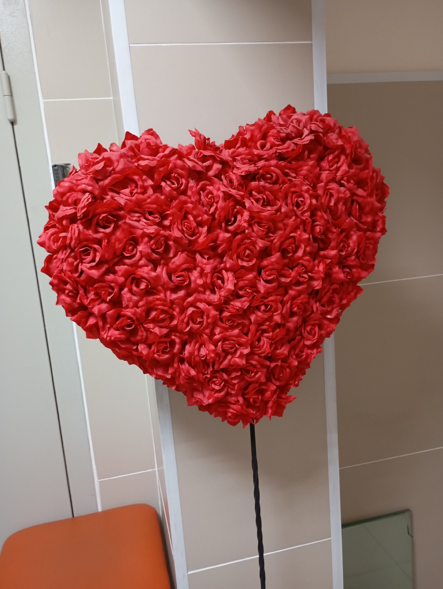 A large, red, heart-shaped decoration made of artificial roses is displayed indoors, possibly in a public space like a store or event venue in Novosibirsk, Russia. The decoration is mounted on a dark, thin pole. The background consists of neutral-colored tiled walls and a light-colored door frame. An orange upholstered seat is partially visible in the lower left corner of the frame.