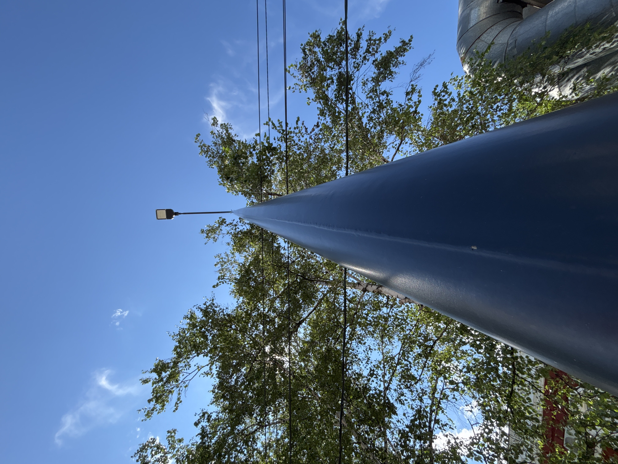 The image depicts a low-angle view of a large, dark blue conical structure in the foreground, pointing upwards towards a clear blue sky. A street lamp with a rectangular fixture is visible at the very top of the conical structure. Behind the structure, lush green trees with visible branches and leaves are present. Several dark electrical wires are strung horizontally across the frame, passing in front of the trees. In the lower right portion of the image, a large, silver-colored pipe with visible seams and banding is present, partially obscured by the conical structure. The overall scene suggests an outdoor urban or industrial setting. The presence of trees and electrical wires indicates an area with some vegetation and infrastructure. No people or animals are visible in the image. The lighting suggests it is daytime with bright sunlight.