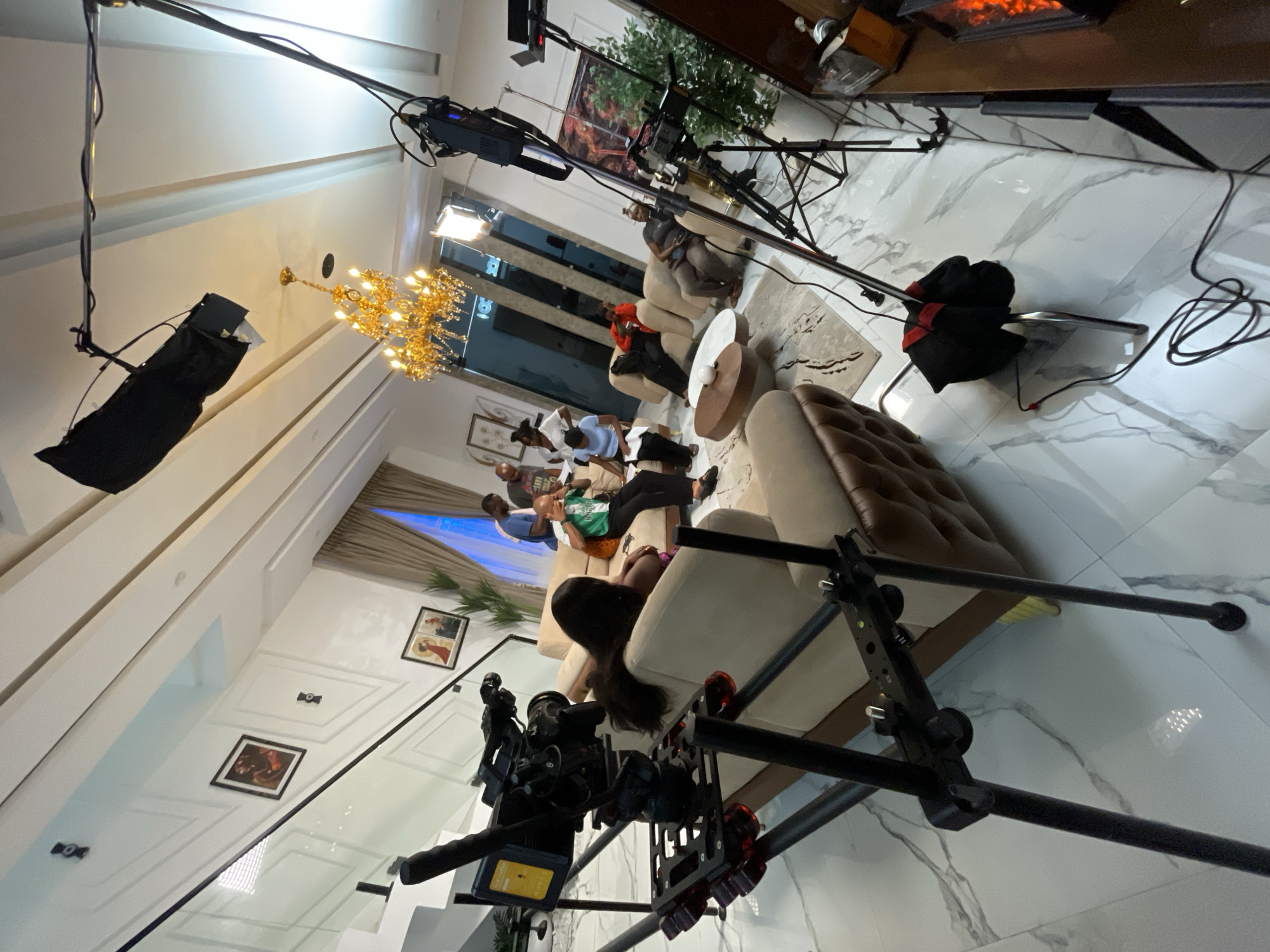 The image depicts an indoor filming scene set in a modern, luxurious apartment. The location is characterized by white marble flooring, high ceilings, and elegant interior design. A large, ornate golden chandelier hangs from the ceiling, providing illumination. Several people are present, appearing to be involved in a film or television production.
In the foreground, a professional video camera is mounted on a complex rig, suggesting a dynamic filming setup. To the left, a staircase with a glass railing leads to an upper level, adorned with framed artwork on the wall.
In the midground, a group of individuals are seated on plush, light-colored sofas. Some are engaged in conversation, while others appear to be reading scripts or preparing for a scene. One person is wearing a green and white shirt and is seated on the sofa, holding a tablet. Another person in a blue t-shirt is standing and looking at something in their hand.
The background reveals more of the apartment's interior, including large windows with beige curtains, potted plants, and additional seating arrangements. The overall atmosphere suggests a professional production underway in a high-end residential setting. The scene is well-lit, with artificial lights, including softboxes, contributing to the professional lighting environment.