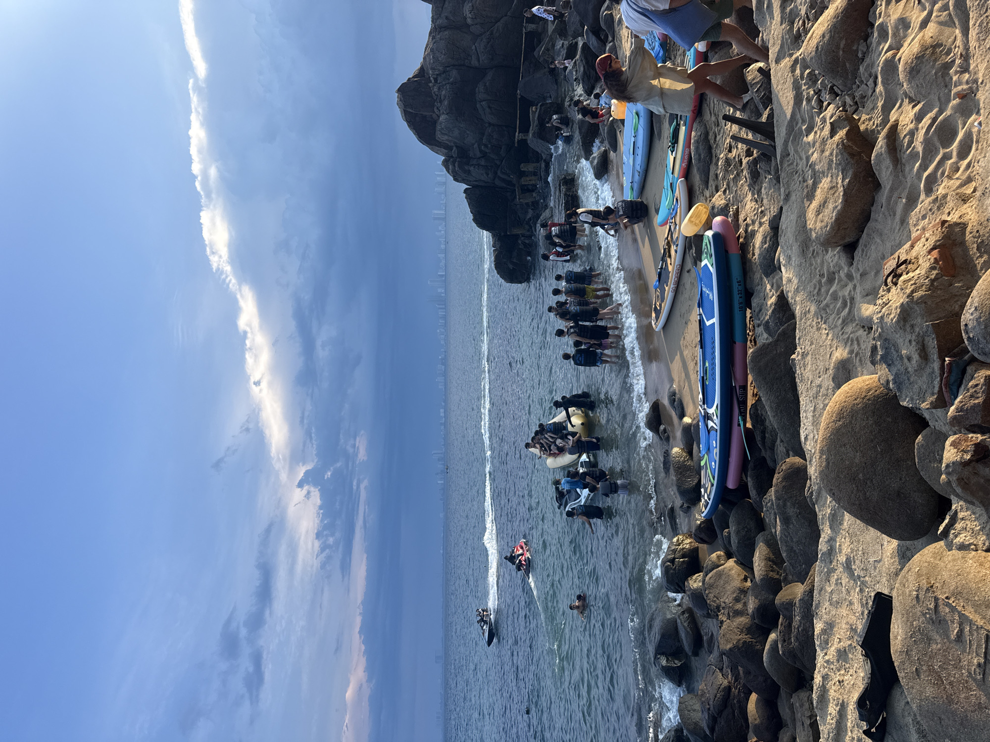 The image depicts a beach scene. Along the shoreline, a group of people are standing in shallow water. Some individuals are engaged in water activities, with at least two jet skis visible in the water. A larger group of people can be seen onshore, positioned between the water and a rocky cliff face. Several stand-up paddleboards lean against the rocky terrain. The scene is set against a backdrop of a partially cloudy sky. In the distance, a city skyline is visible across the water. The image appears to be taken from a high angle, looking down onto the beach.