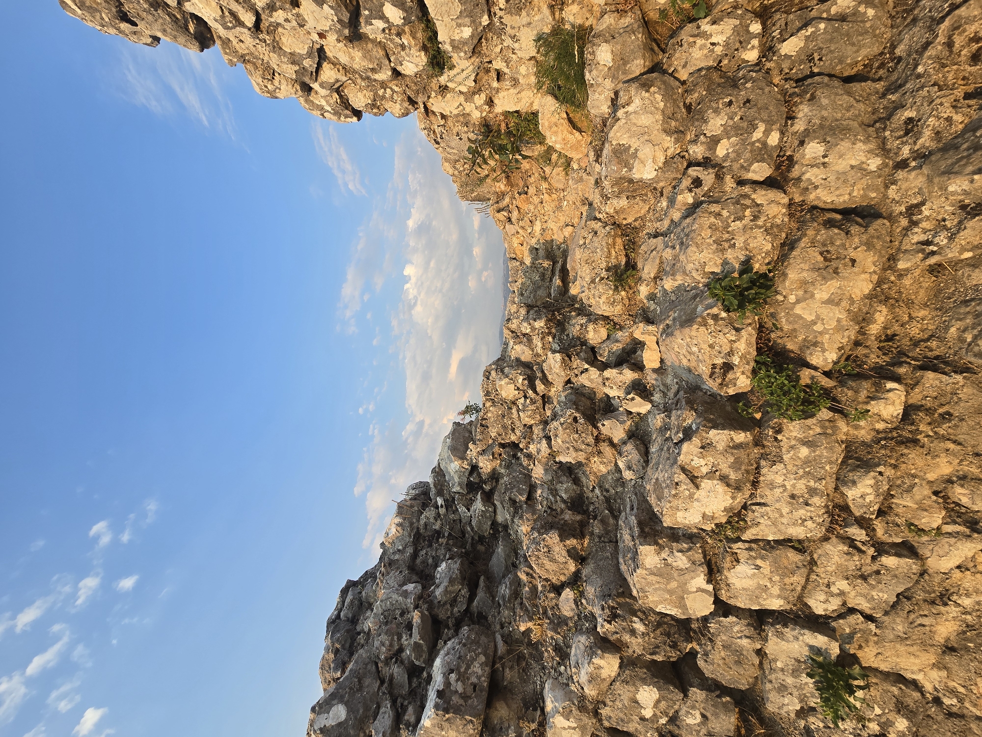 The image shows the ruins of a stone fortress in Medun, Montenegro, under a bright blue sky with scattered white clouds. The foreground is dominated by large, rough-hewn stones that form the crumbling walls of the structure. Some of these stones are lit by the warm glow of the sun, casting shadows and highlighting their textures. Small green plants are growing in the crevices between the stones, indicating the passage of time and the natural reclamation of the ruins. In the distance, a hazy landscape can be seen, suggesting a mountainous terrain. The overall impression is one of ancient history and enduring natural beauty.