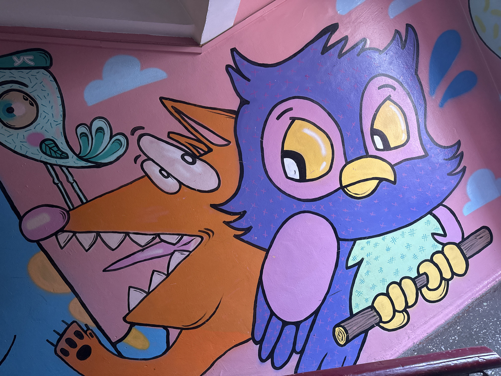 This image is a close-up shot of a vibrant, cartoon-style mural painted on a pink surface. The mural features several whimsical characters. In the foreground, a large, purple owl-like creature with big yellow eyes and a yellow beak is depicted. It has a purple body adorned with small pink crosshatch marks and a light green chest area with a subtle pattern. The owl is holding a brown stick with both hands, which are rendered in yellow. Behind and above the owl, a portion of an orange creature, possibly a fox or dog, is visible. This creature has sharp teeth and a large, pink mouth. Above these characters, a turquoise fish with a single eye and a green tail fin is drawn. The background consists of a light blue sky with small, light blue cloud shapes. The mural appears to be located on an interior surface, possibly a wall or a bench, within the city of Biysk, Russia, though a specific location cannot be pinpointed from the image alone.