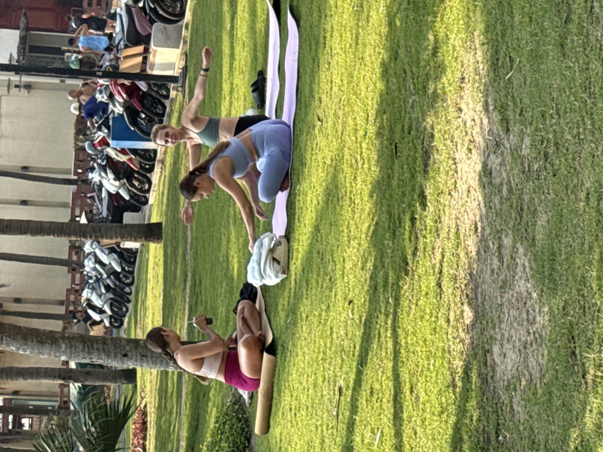 Captured in Đà Nẵng, Vietnam, the scene depicts three women engaged in outdoor activities on a grassy lawn near a street. Two women are practicing yoga on yoga mats, while the third woman is sitting on her mat, possibly using her phone. The location is adjacent to a street lined with parked motorbikes and palm trees. The overall environment suggests a public park or recreational area in the city.