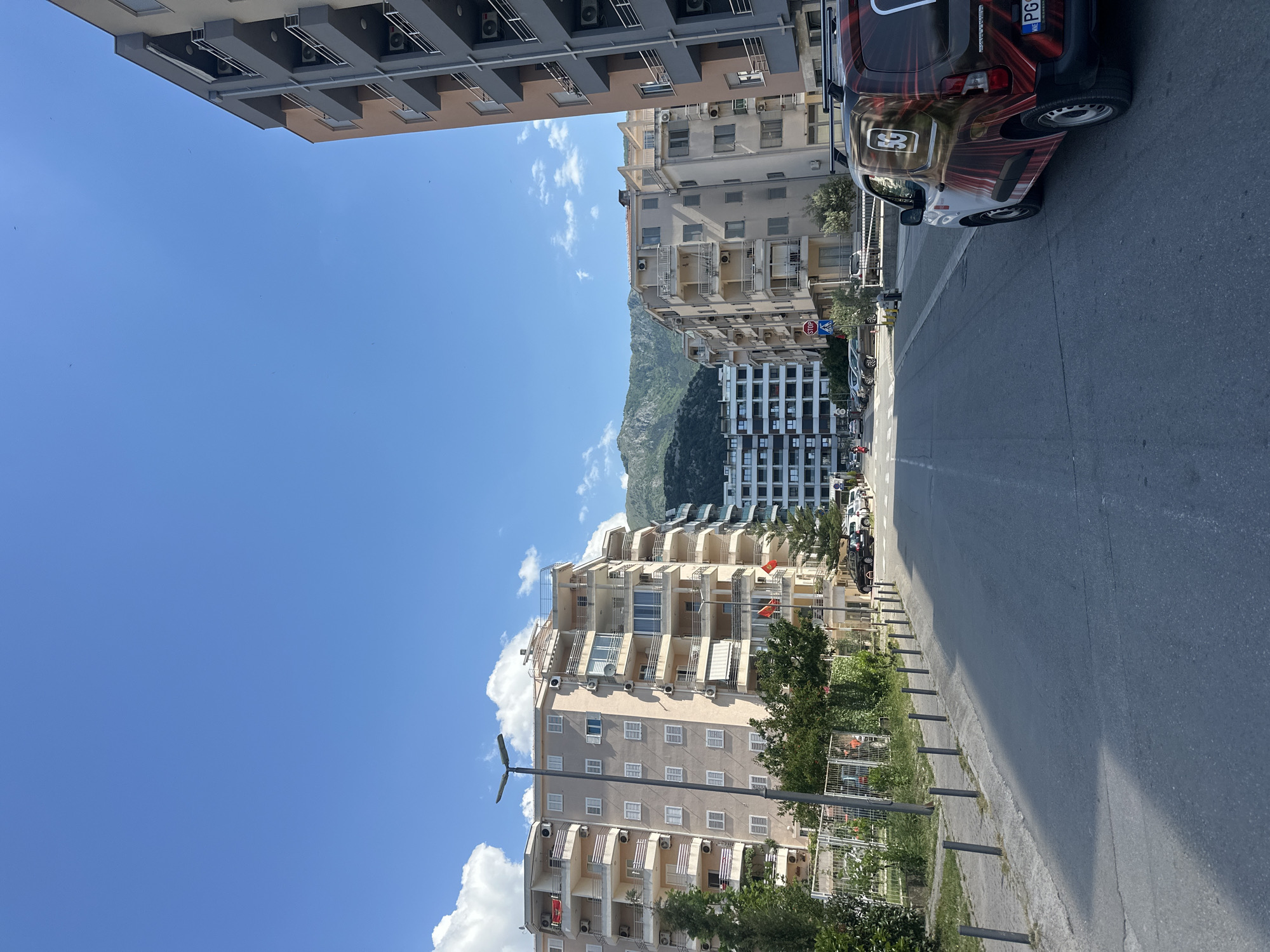 The image depicts a street scene in Budva, Montenegro. The perspective is from a low angle looking down a straight road lined with apartment buildings on both sides. The buildings are multi-story with beige or gray facades and balconies. A mountain range is visible in the distance between the buildings. A car is parked on the right side of the street. The road is paved and marked with a solid white line. There are trees and fencing along the sidewalk. The sky is blue with scattered white clouds.
