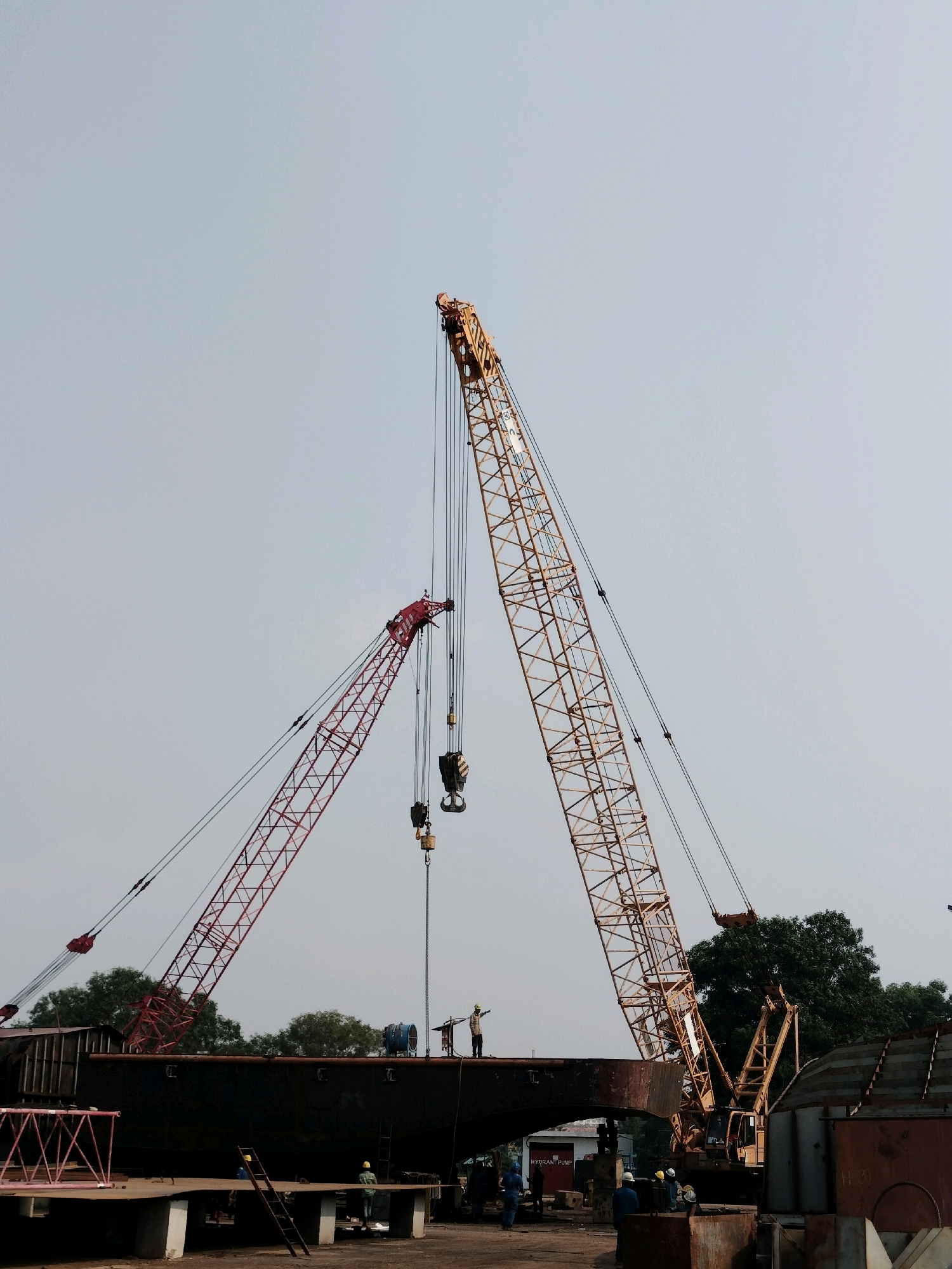 This image depicts a shipyard scene in Batam, Indonesia, likely in the Kabil or Batu Ampar industrial areas, characterized by the presence of large industrial cranes and shipbuilding activities.
The foreground shows a large, dark-hulled ship section under construction, resting on a platform supported by concrete pillars. A red metal scaffolding structure is visible to the left. Several workers in hard hats and work clothes are present on and around the ship's deck and on the surrounding ground. A ladder leans against the ship's hull.
Dominating the midground and background are two large cranes. A red lattice-boom crane is angled upwards on the left, with multiple cables and pulleys. A larger, yellow lattice-boom crane stands on the right, also angled upwards, with its boom extending prominently into the sky. This crane appears to be in operation, with its hoist line and hook visible.
In the distance, trees line the horizon, suggesting an area with some vegetation bordering the industrial site. The sky is overcast with a pale blue-grey hue. The overall impression is one of active industrial construction, typical of a shipyard. The presence of large marine vessels or their components, specialized heavy machinery like cranes, and workers engaged in construction or repair are all indicative of a shipbuilding or ship repair facility.
