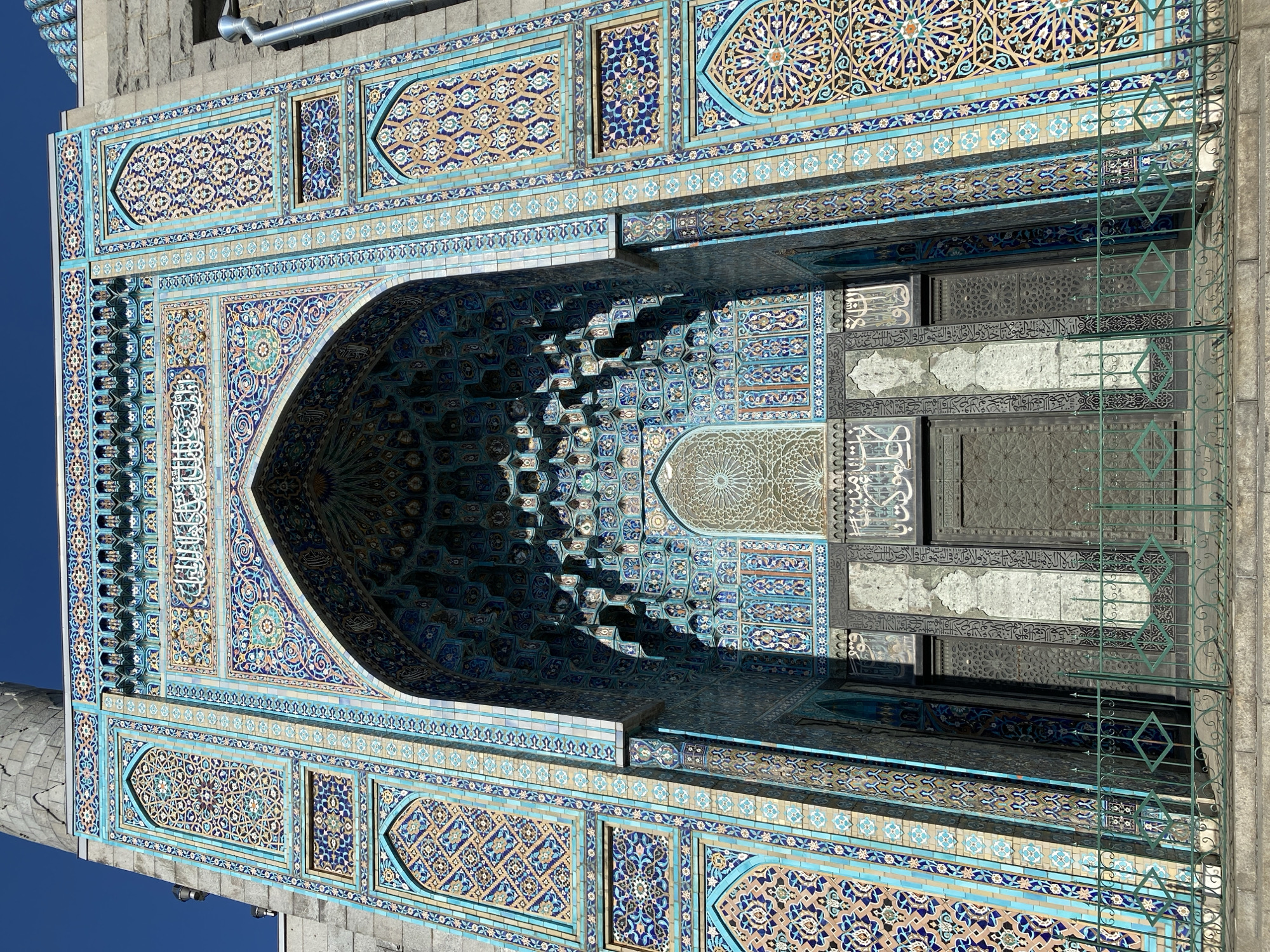 This is a frontal, eye-level, full-frame photograph of the entrance of the Saint Petersburg Mosque, located in Saint Petersburg, Russia. The mosque's facade is adorned with intricate blue, gold, and white tilework featuring geometric patterns and Arabic calligraphy. The central archway of the entrance is deeply recessed and decorated with a muqarnas (honeycomb vaulting) ceiling. Two large, dark metal doors are visible beneath the arch. A green metal fence is positioned in front of the mosque, with a stone staircase leading up to the entrance. The sky above is clear and blue, with the stone minaret of the mosque visible on the left side of the frame. The photograph is taken on a sunny day, casting shadows on the intricate facade. There are no people visible in the image.