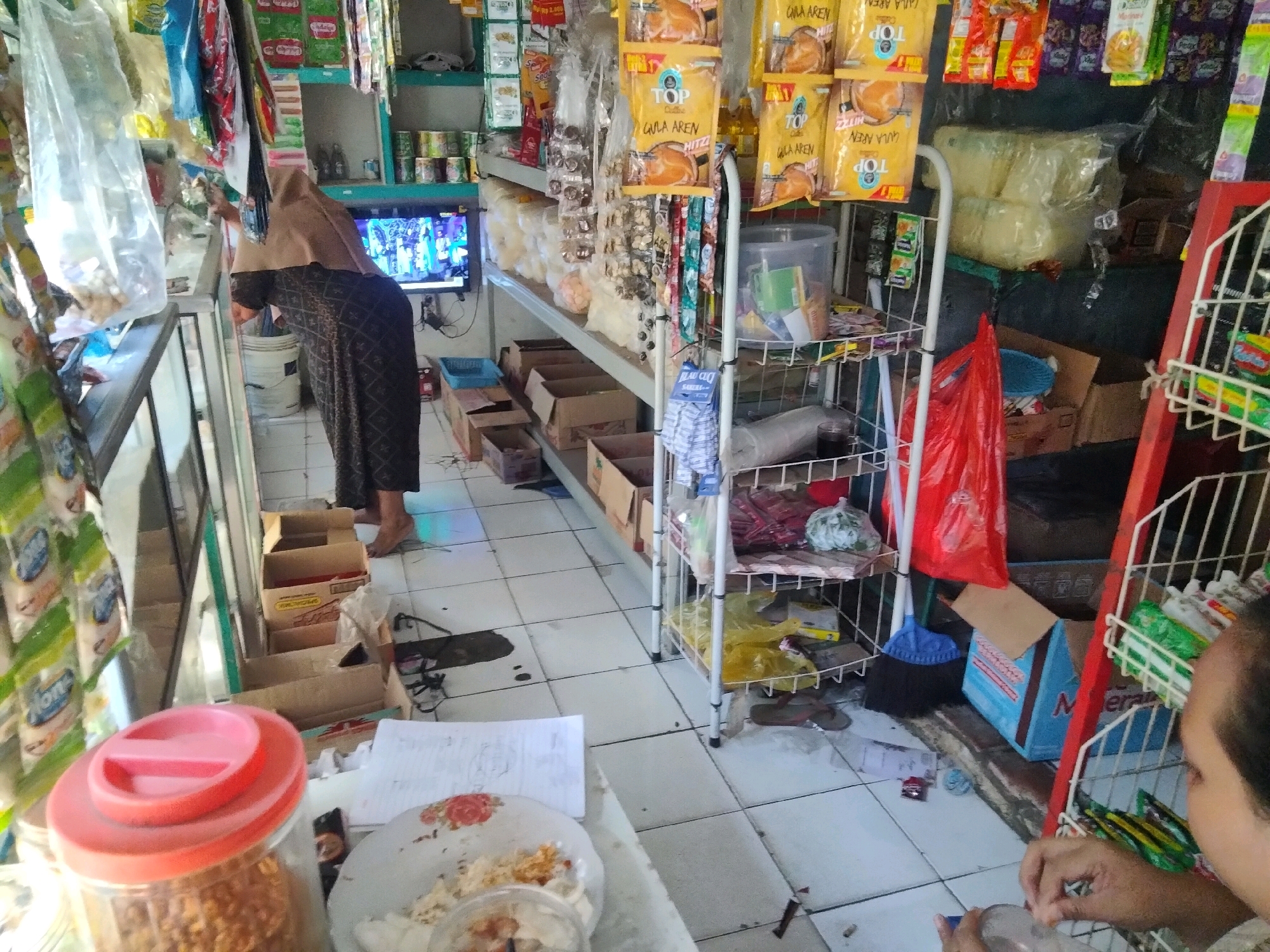The image depicts the interior of a small convenience store, possibly a warung, located in Suka Sari, Indonesia. The store is cluttered with various goods displayed on shelves and hanging from the ceiling. In the foreground, a person is visible from the shoulders up, their hands engaged with a bottle. Behind them, a woman wearing a hijab and a patterned dress is bent over, facing a television screen that is displaying a bright, colorful image, likely a program or advertisement.
The store's shelves are stocked with packaged snacks, drinks, and household items. Cardboard boxes are stacked on the floor and in various corners. A red shelving unit with white wire racks stands on the right side of the image, displaying more products. A broom and a blue plastic bucket are visible near the shelving unit. The floor is tiled. The overall impression is of a busy, small retail establishment.
