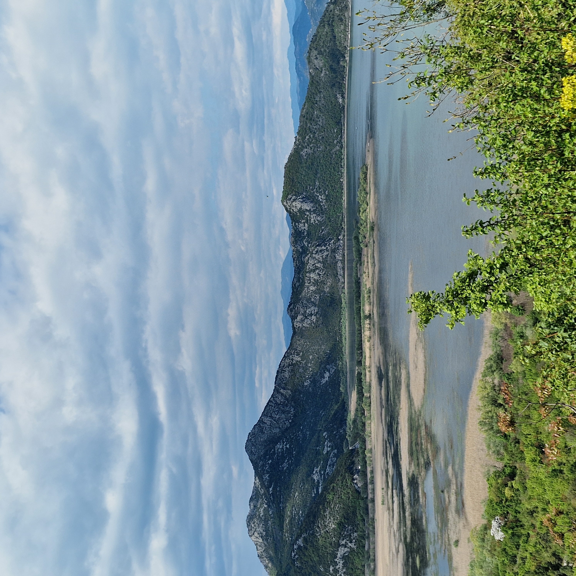 The image shows a landscape in Godinje, Montenegro. A lake stretches along the right side of the frame, next to a grassy and sandy shoreline. Green foliage grows densely on the right edge of the image. A mountain range runs parallel to the lake on the left, with a sky of pale blue and streaky white clouds in the background.